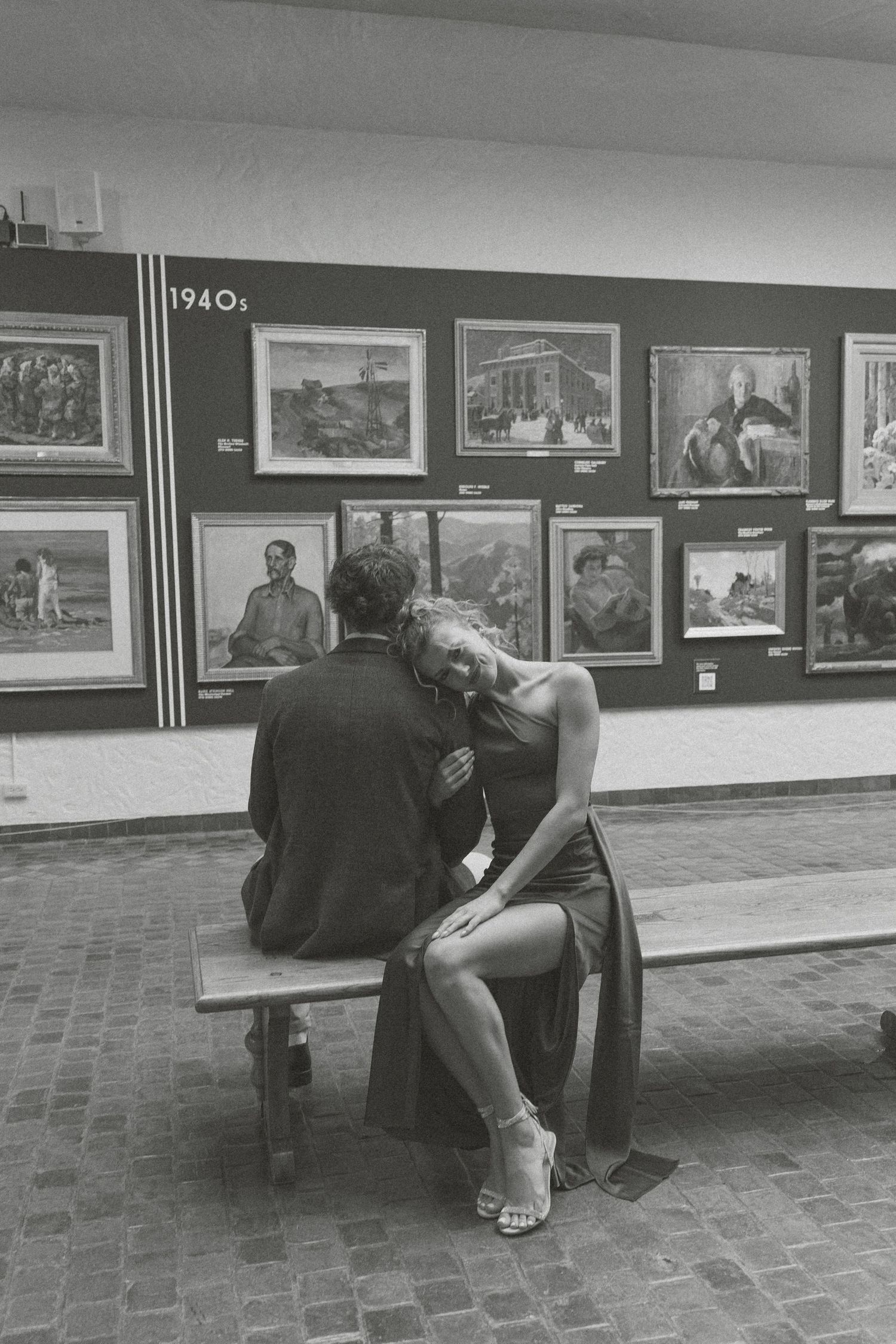 A black and white photograph shows two people sitting on a bench in an art gallery with framed artwork on the walls.