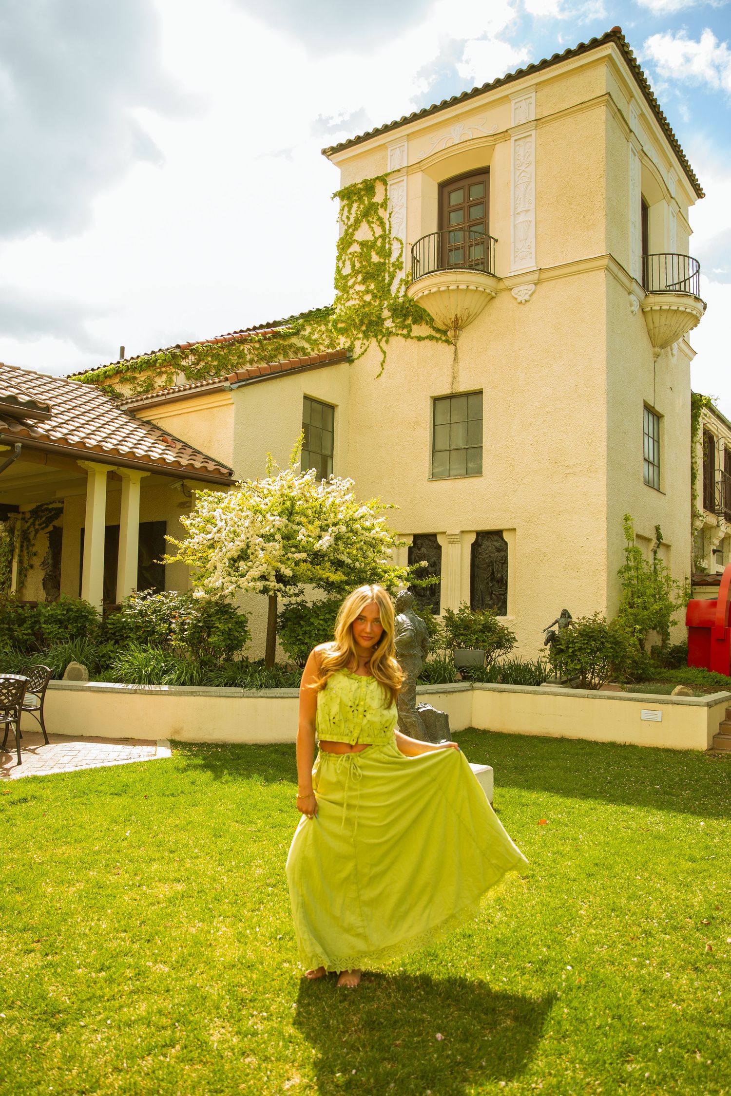 Woman in flowing yellow-green maxi dress twirling on green lawn in front of elegant Mediterranean-style villa on sunny day.