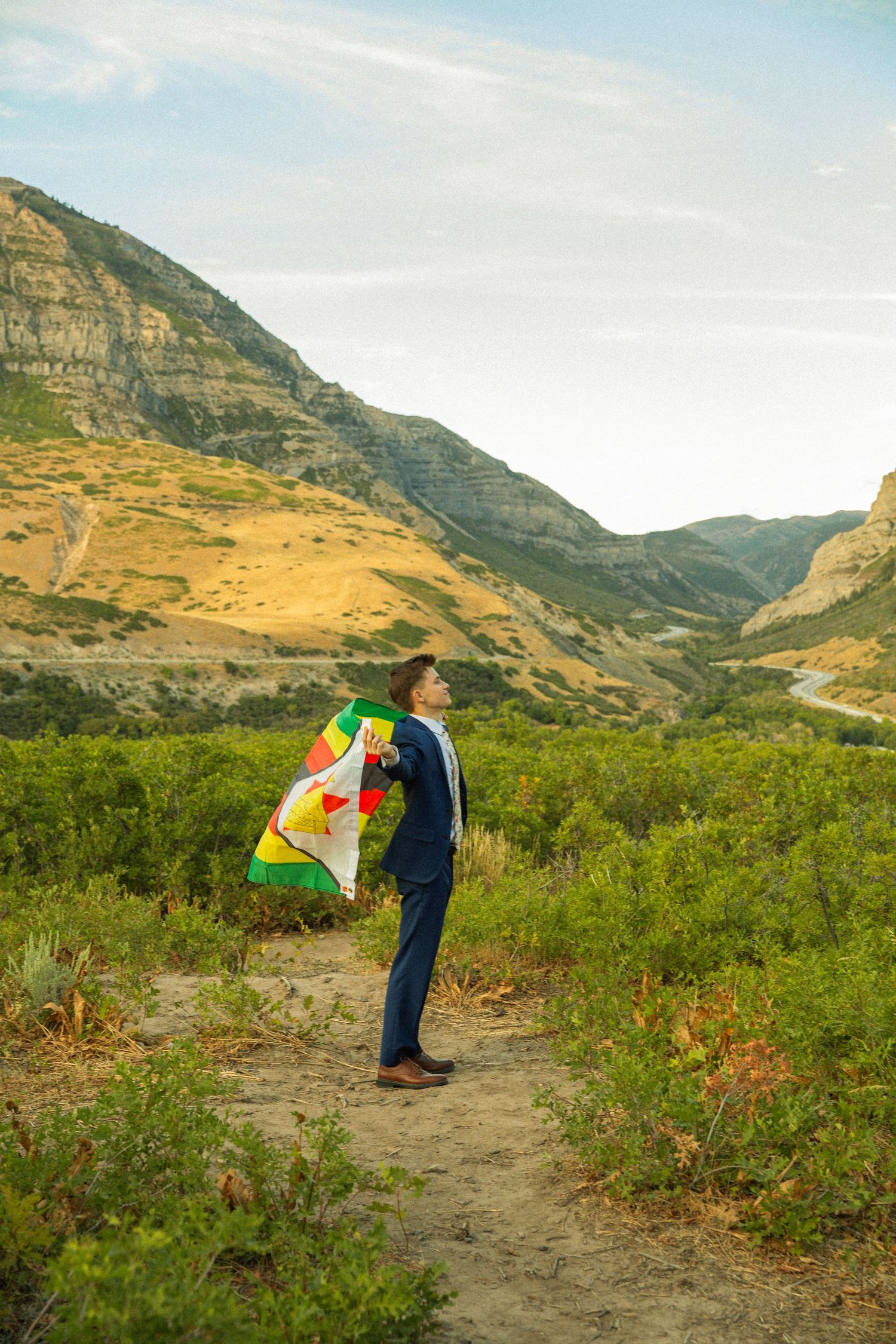 Person bending backwards on a mountain trail surrounded by rugged terrain and golden vegetation under blue skies.