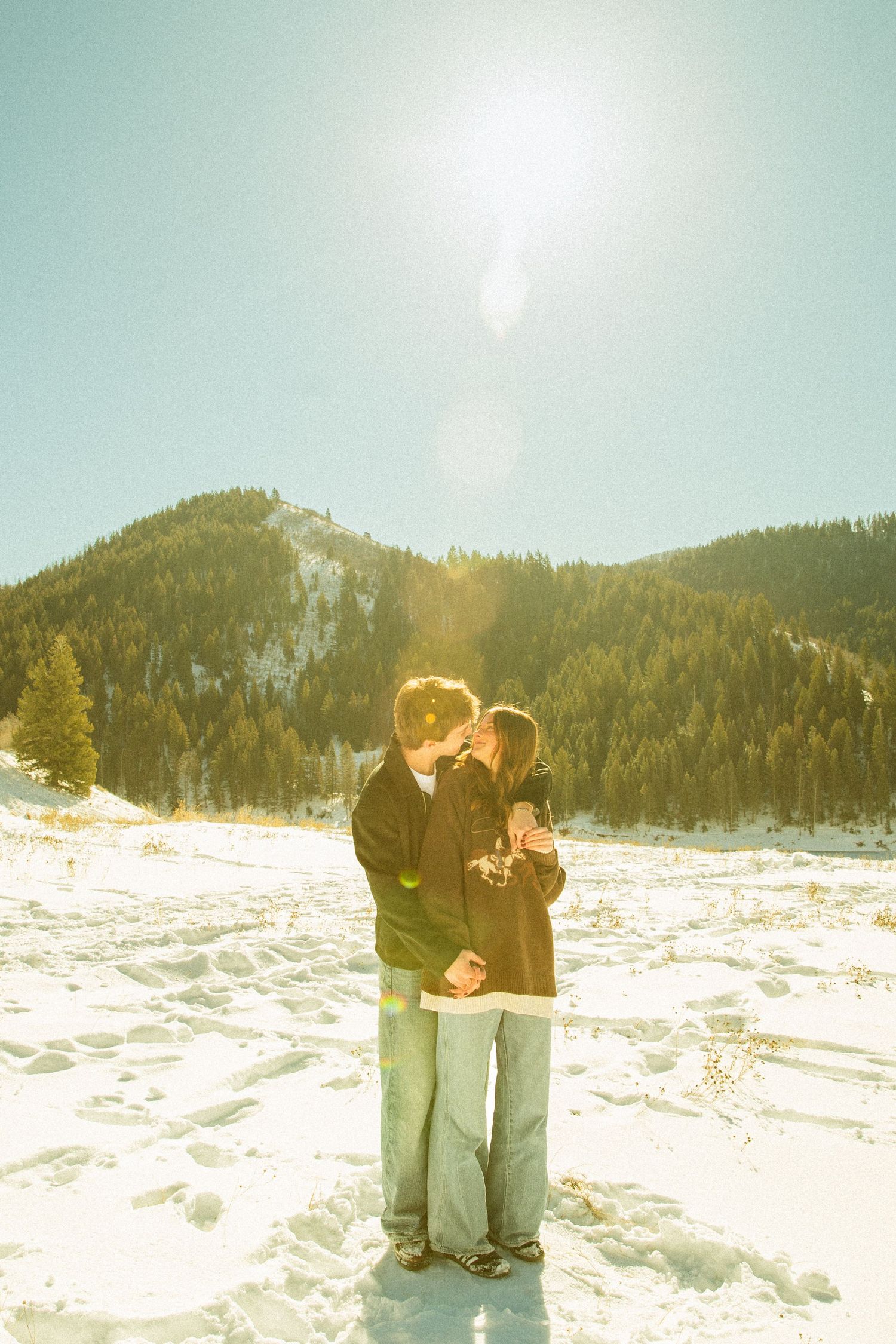 A couple embraces in a snowy winter landscape with sunlit mountains and evergreen trees in the background.