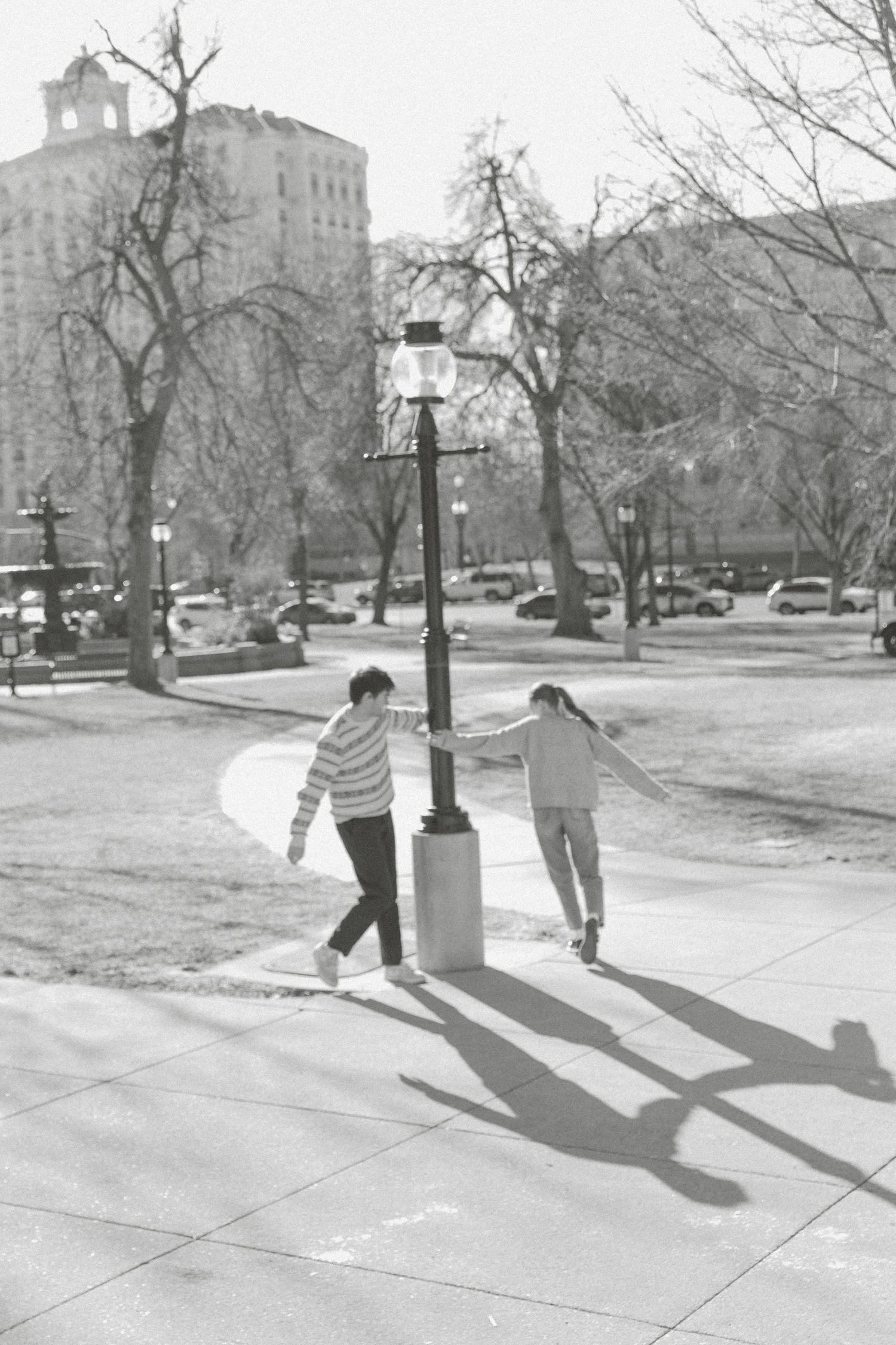 Two children cast long shadows while playing together on a sidewalk in a black and white urban winter scene.
