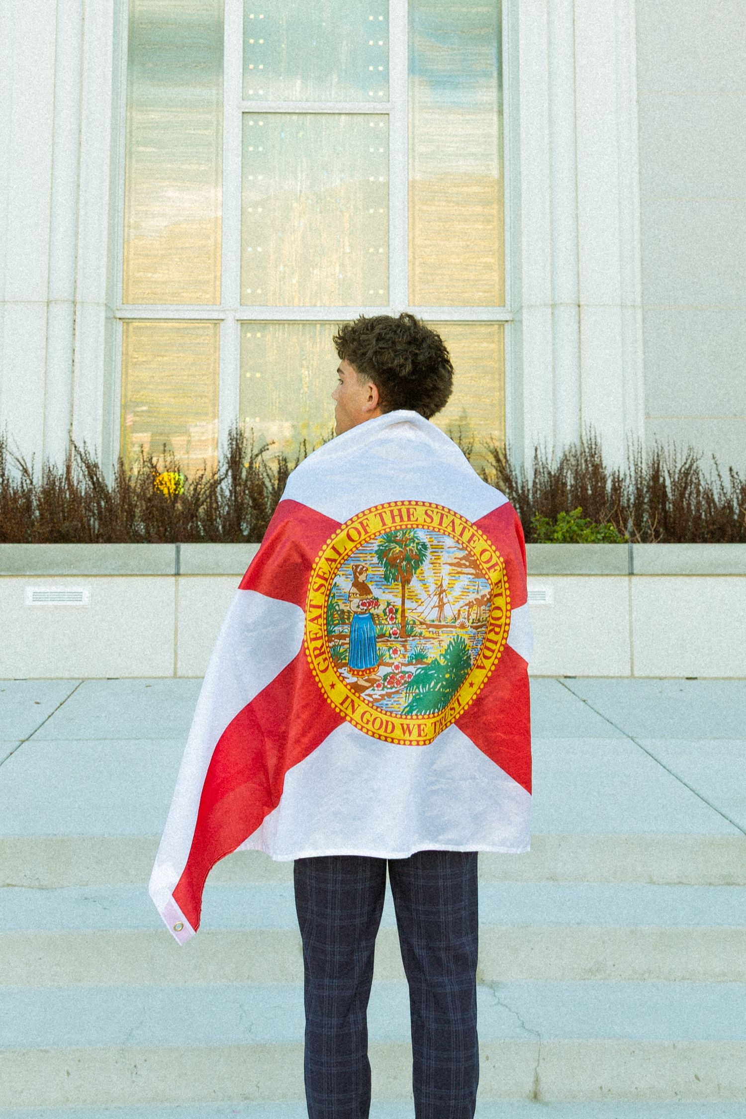Person draped in Florida state flag standing on steps facing a large building window.
