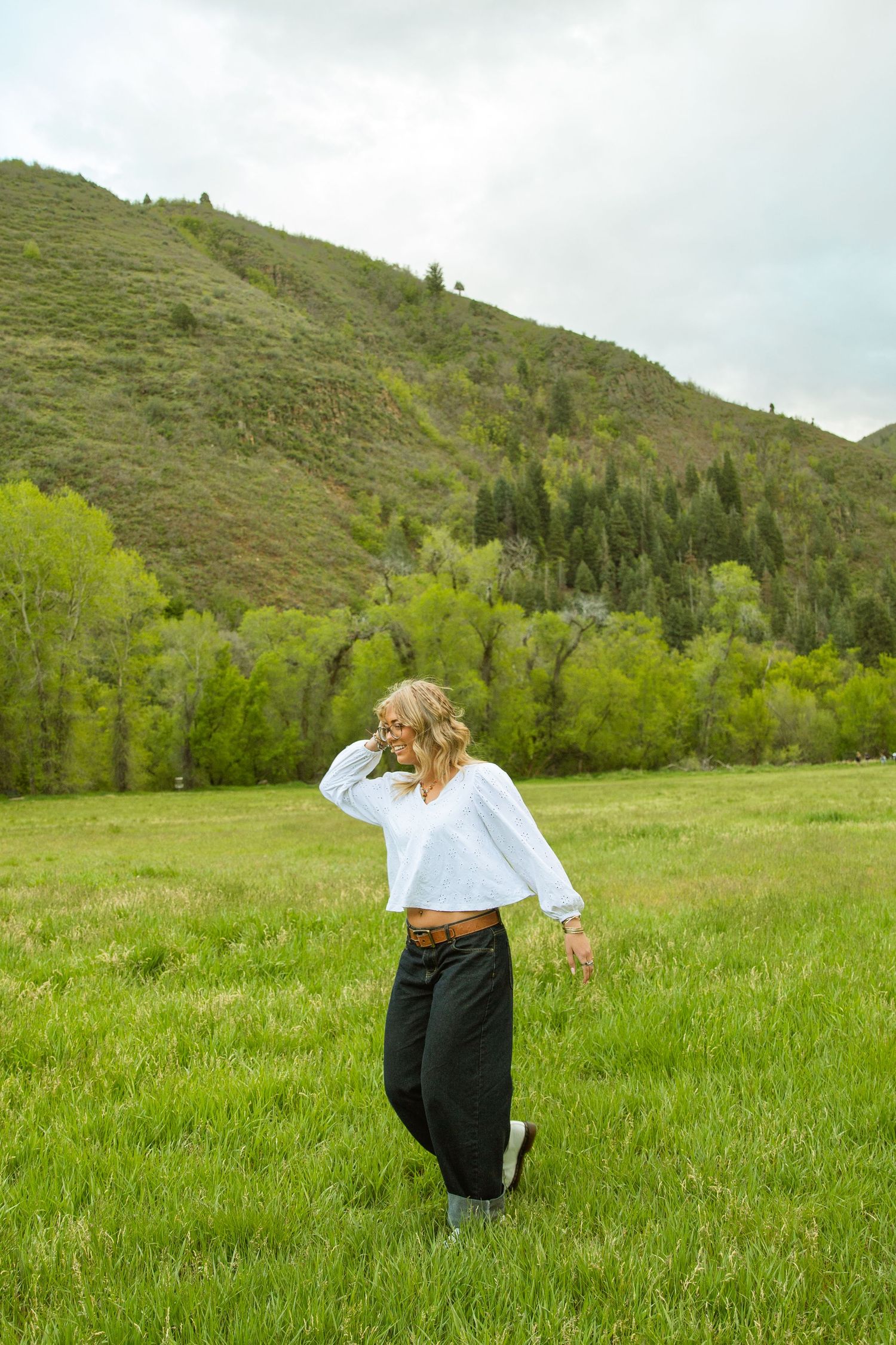Woman in white crop top and black pants walking in a lush green meadow with mountain backdrop.