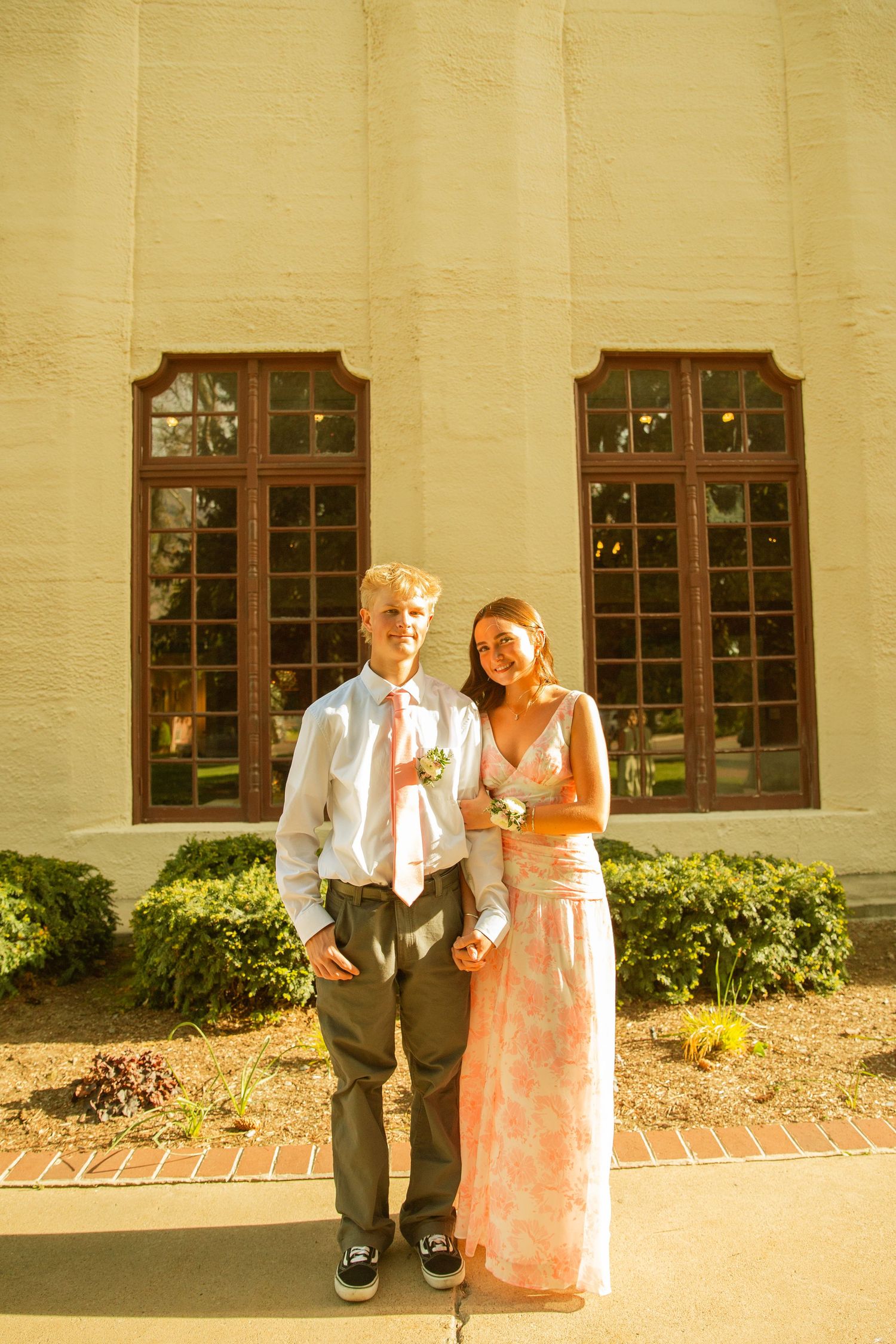 A young couple dressed in formal prom attire poses outside a building with large windows and lush greenery.