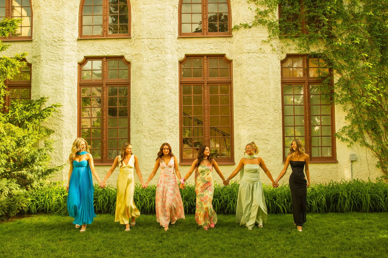 Six women in colorful formal gowns hold hands on a green lawn in front of an elegant white building.