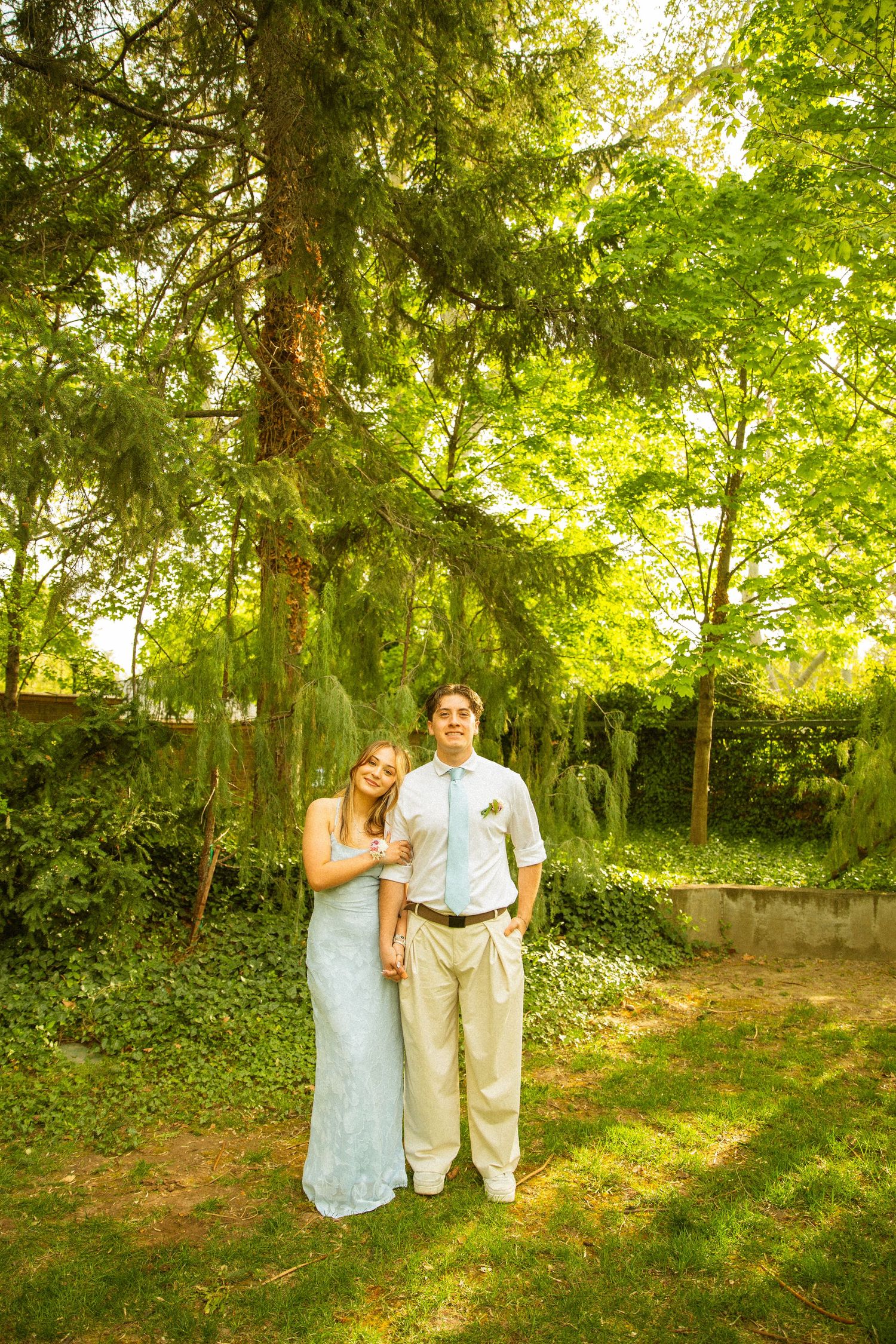 Young couple in formal attire posing together in a lush green garden setting on a sunny day.
