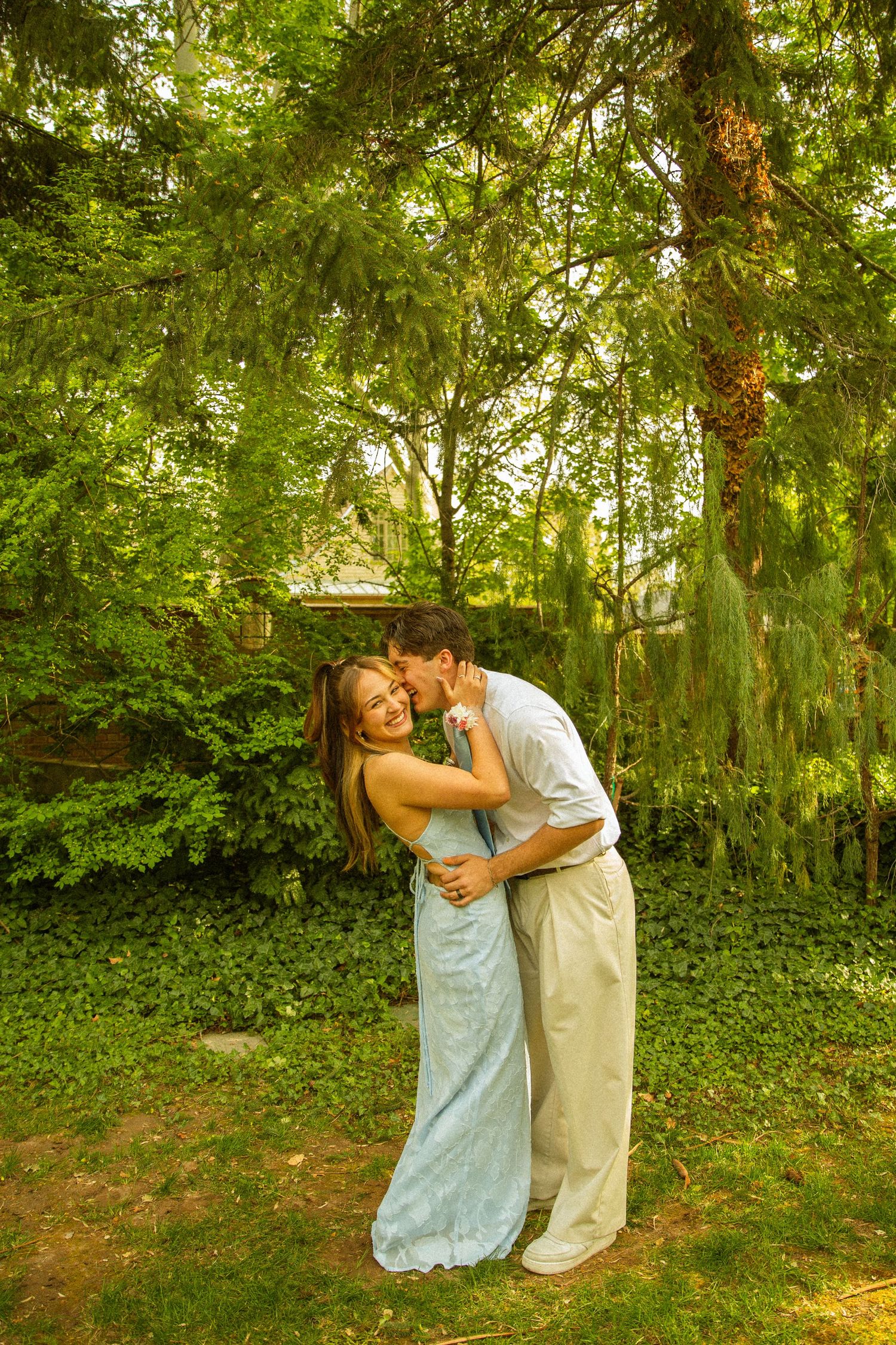 Couple sharing a romantic kiss in a lush green garden, woman in light blue dress smiling joyfully.