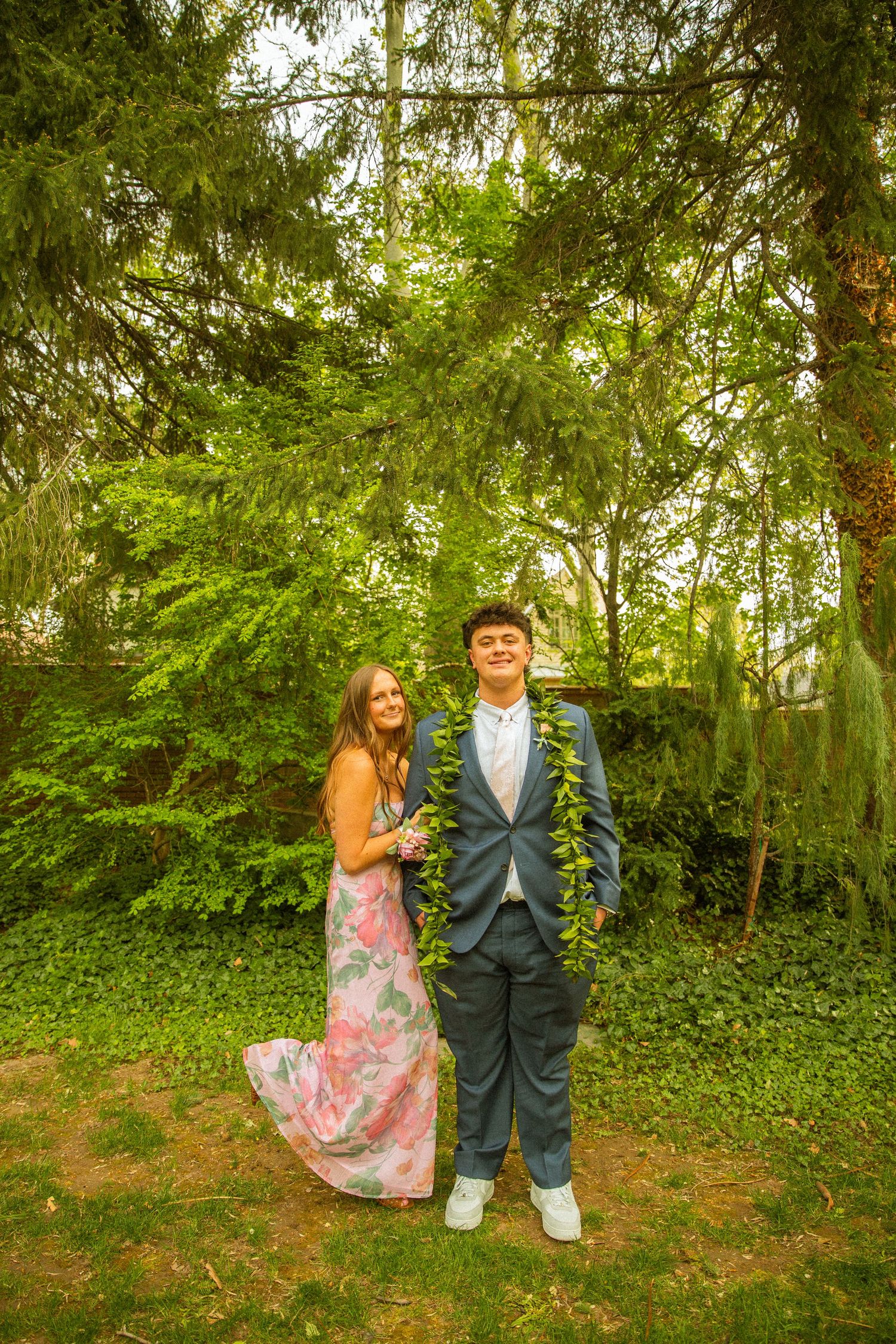 A couple in formal attire poses outdoors, woman in floral dress, man in blue suit with green lei, surrounded by lush trees.
