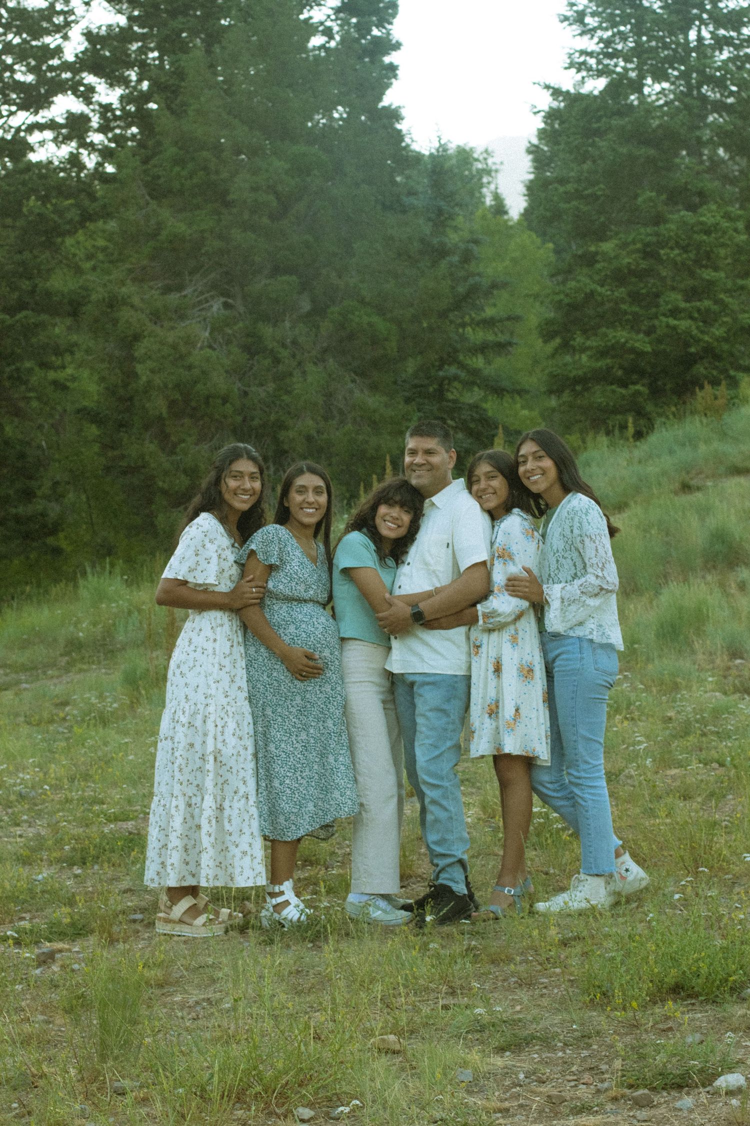 A group of people in casual summer clothing stand together outdoors on a grassy hillside among evergreen trees.