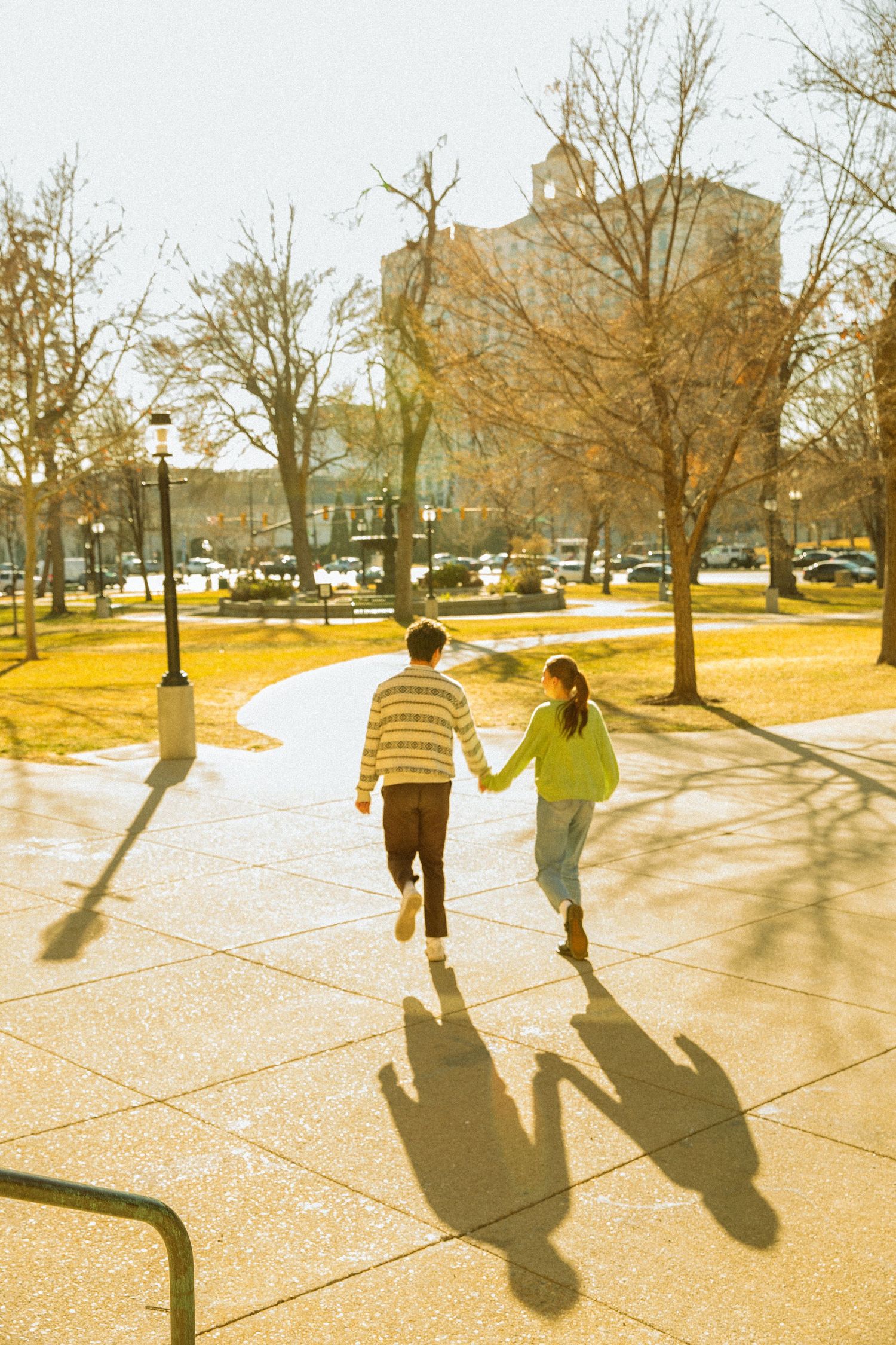 Three people cast long shadows while walking together on a sunlit concrete path through a park on a bright day.