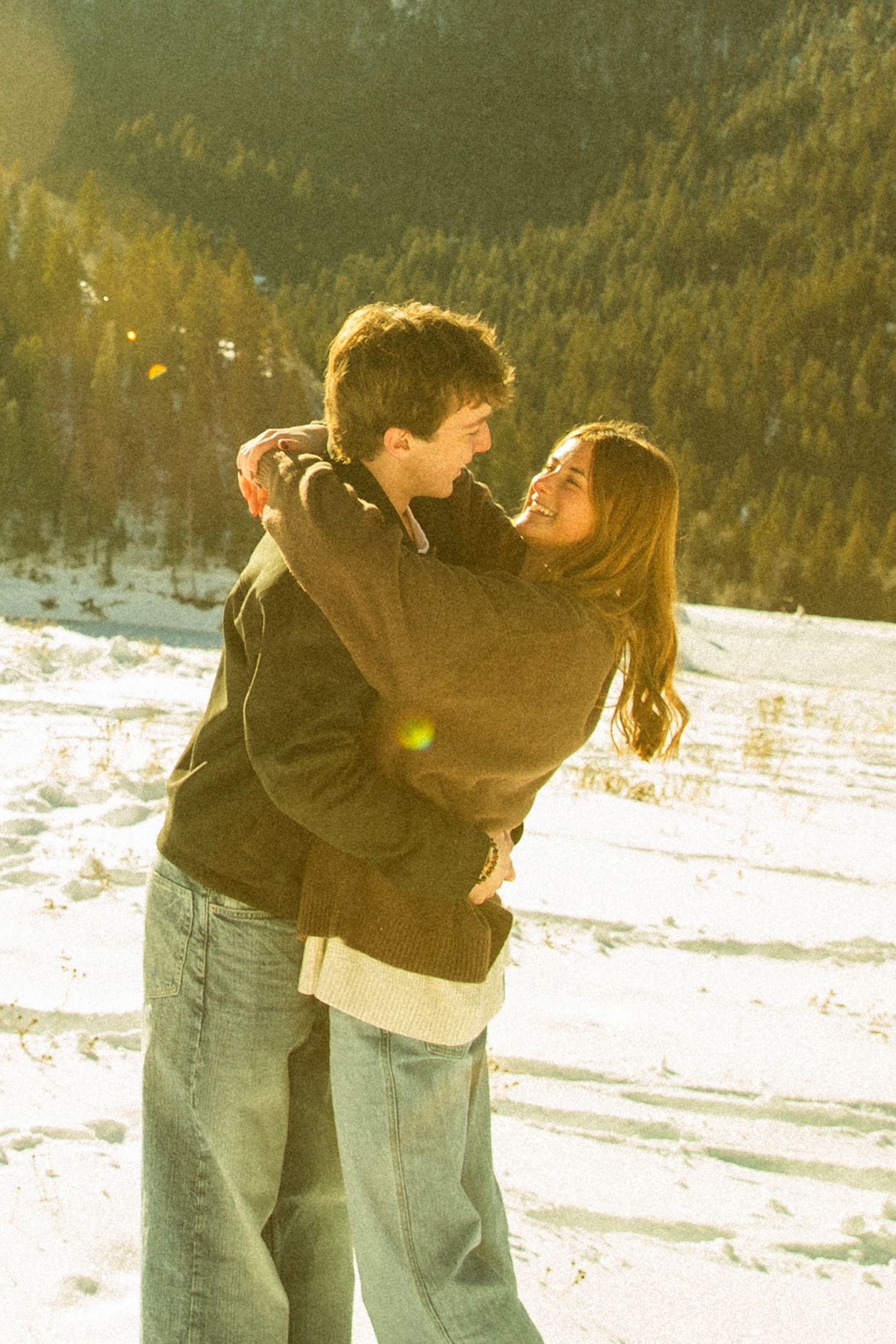 A couple embraces in a snowy winter landscape with sunlight filtering through trees in the background.
