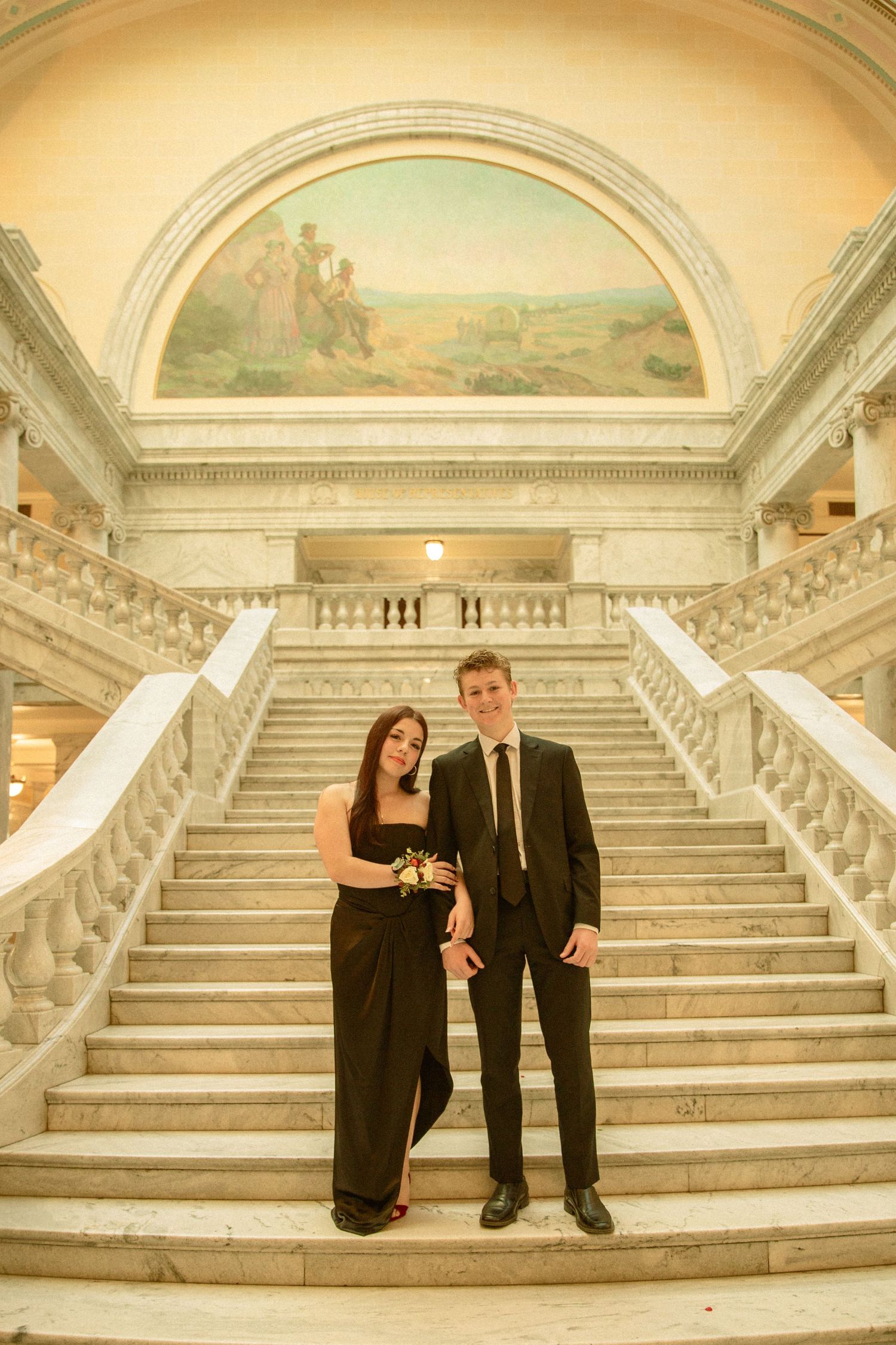 A couple in formal black attire poses on grand marble stairs in an elegant architectural setting.