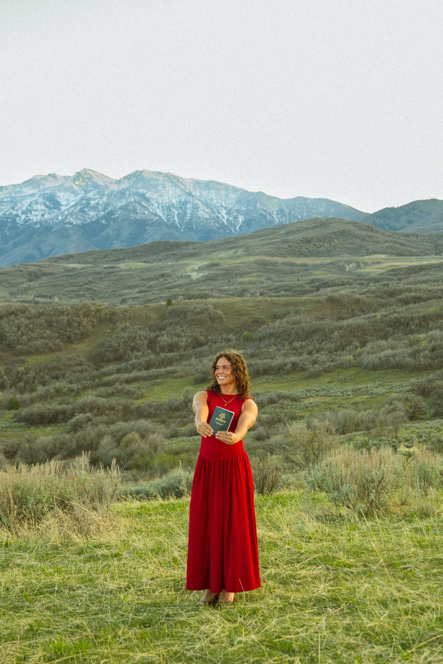 Woman in red dress holding a book in a scenic mountain landscape with green hills and snow-capped peaks.