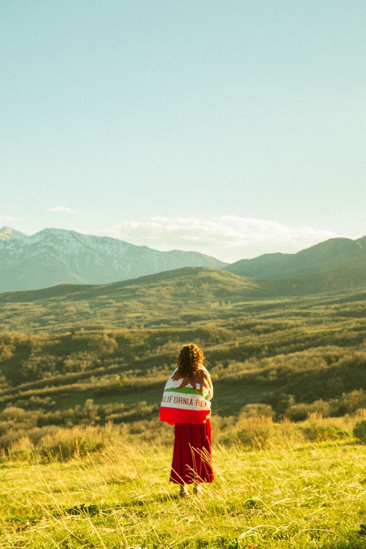 Woman in red skirt draped in a flag stands facing lush green mountains under a bright clear sky.