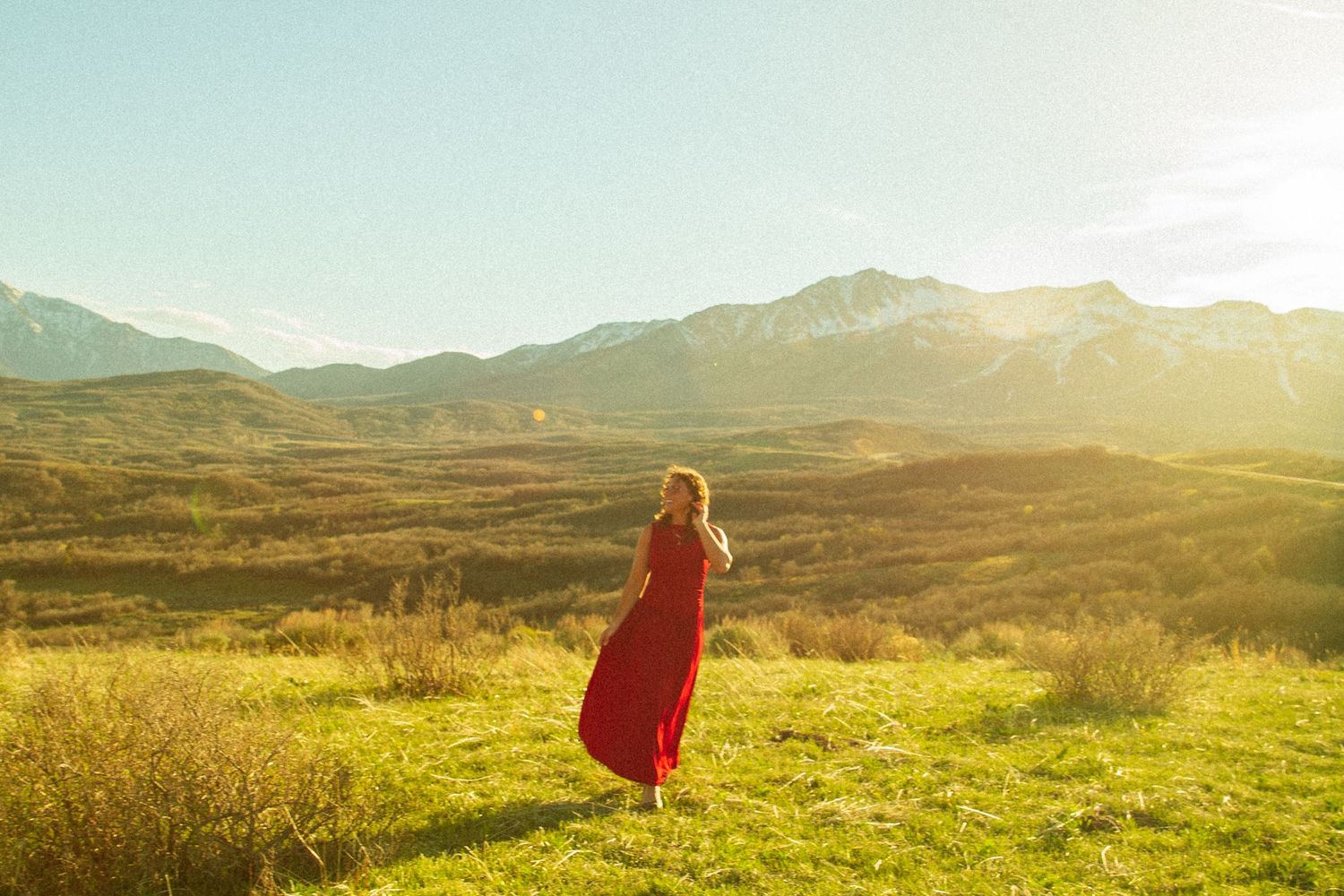 Woman in red dress standing in sunlit mountain valley landscape at golden hour.