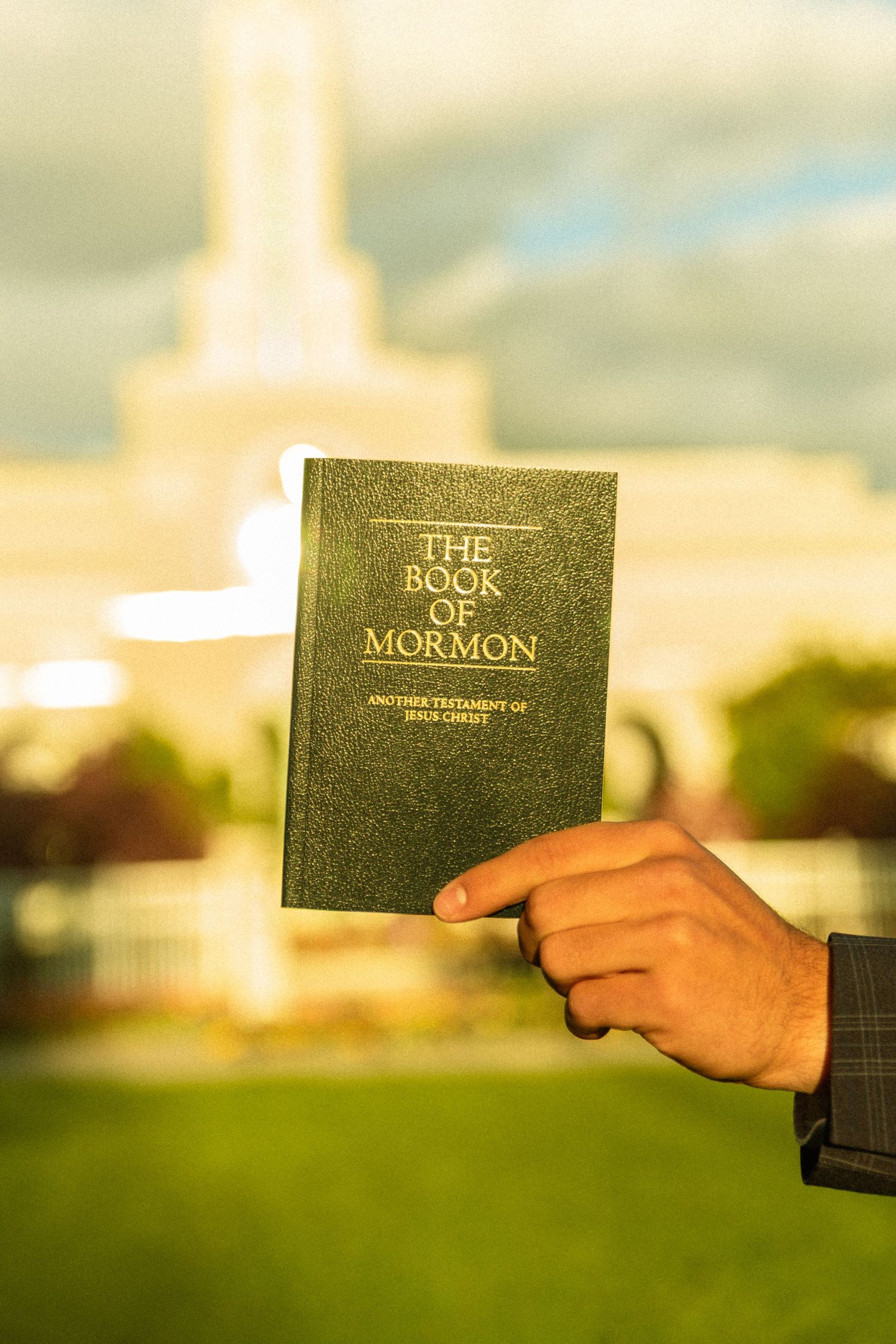 Hand holding The Book of Mormon scripture outside an LDS temple with green grass and cloudy sky background.
