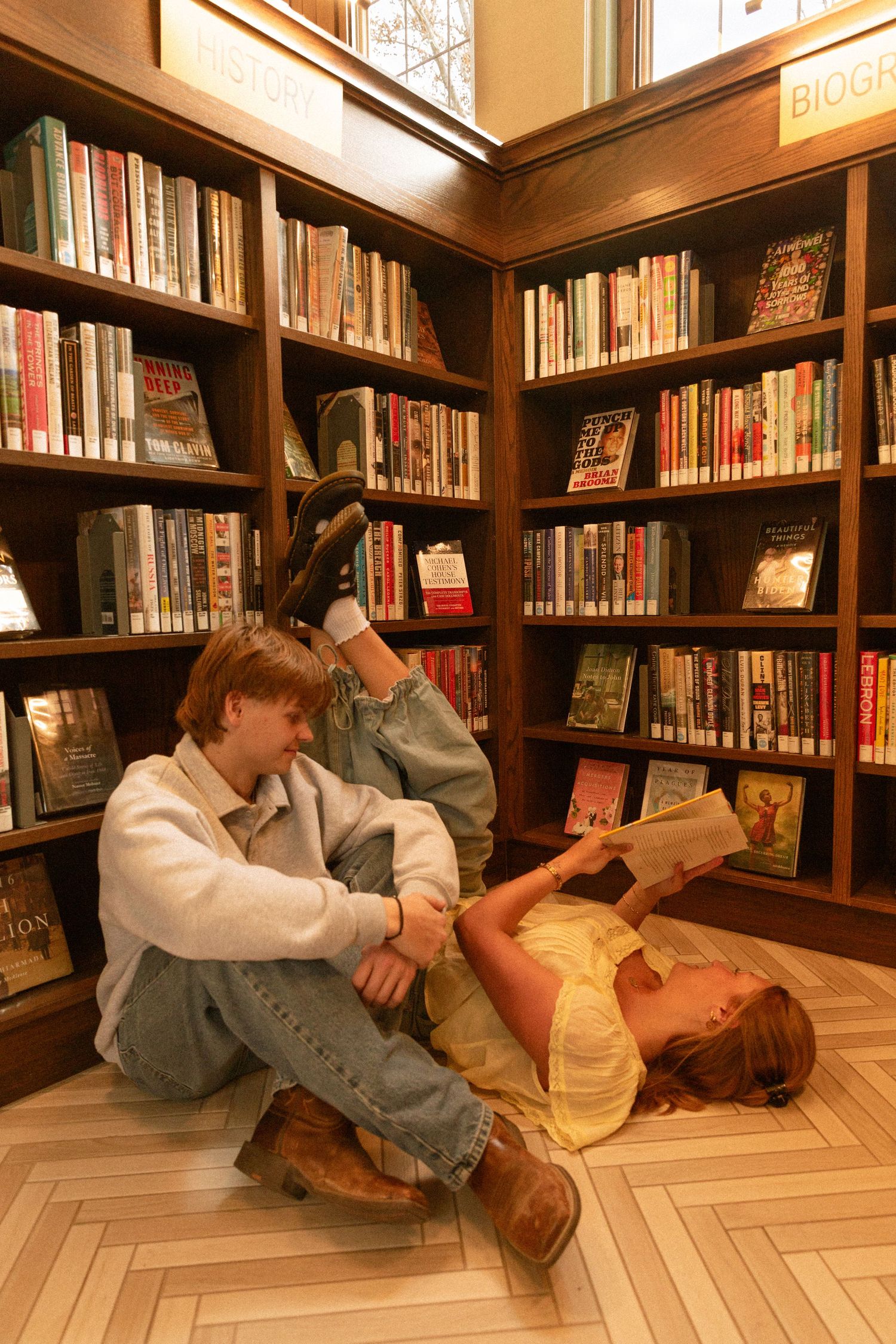 Students relax and read books on the floor of a cozy library corner with wooden bookshelves filled with literature.