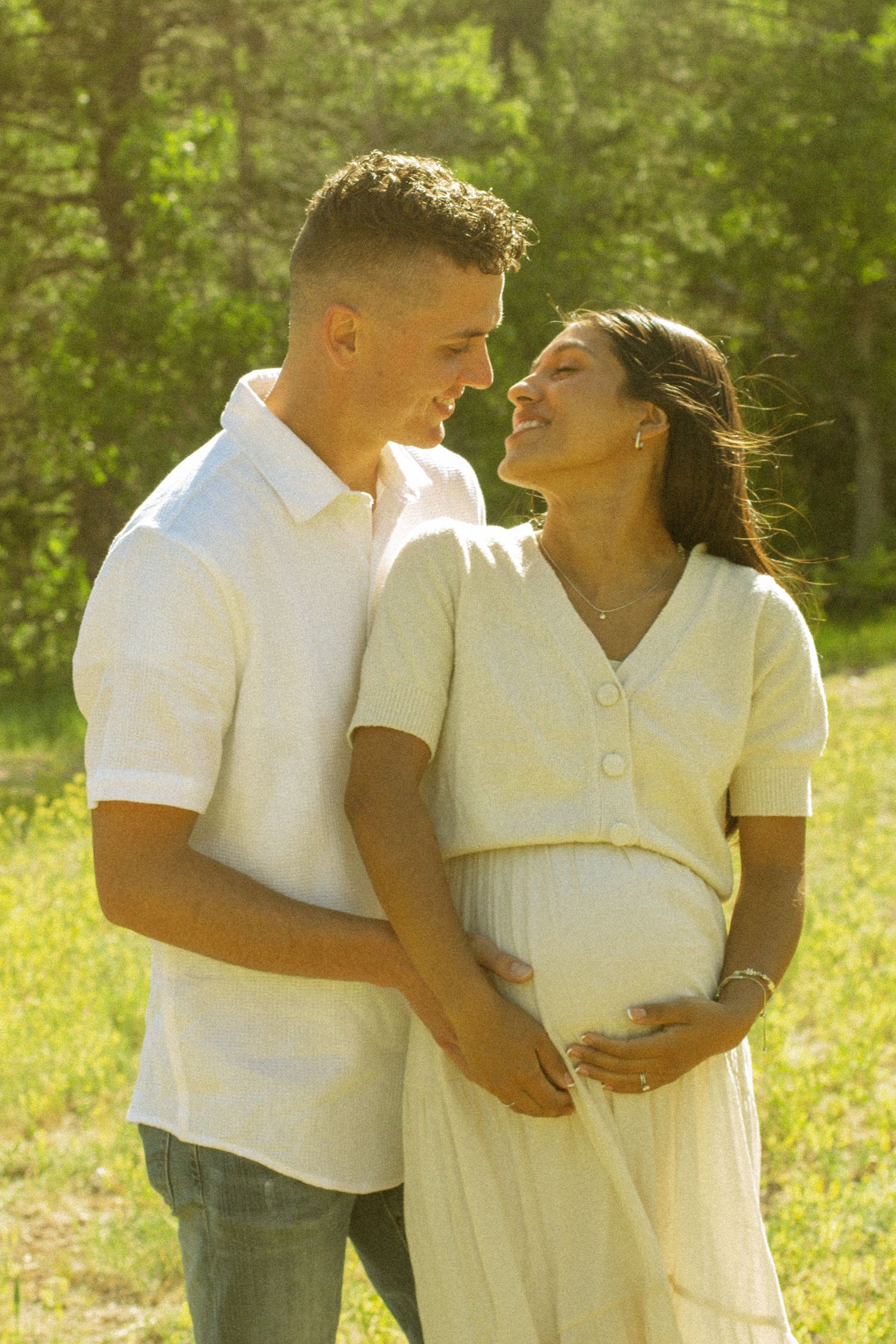 A romantic maternity photo of a couple in white outfits embracing in a sunlit outdoor setting.