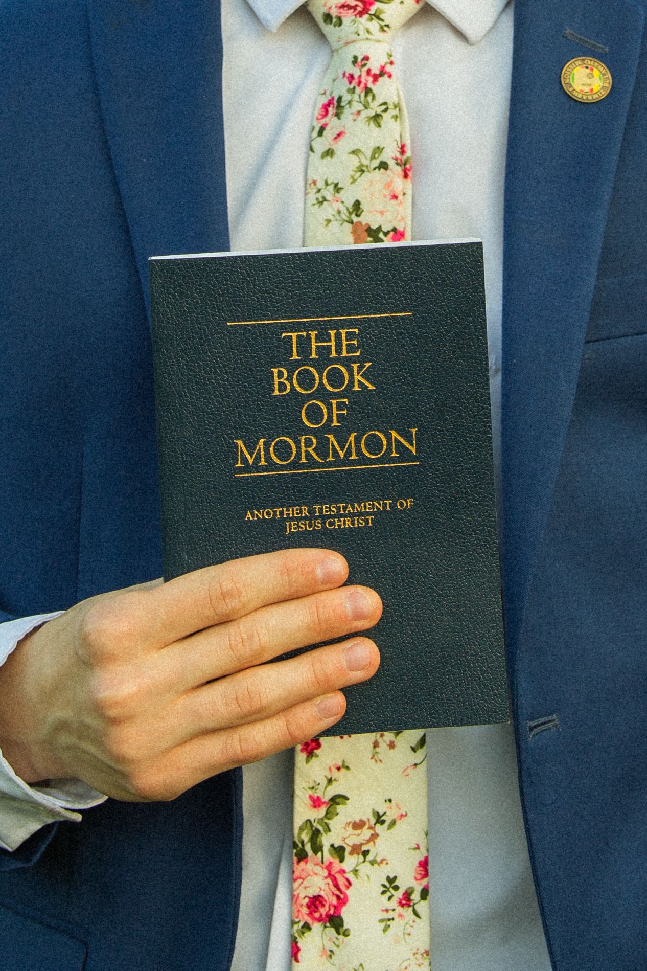 Close-up view of hands holding a navy blue Book of Mormon against a floral patterned tie.