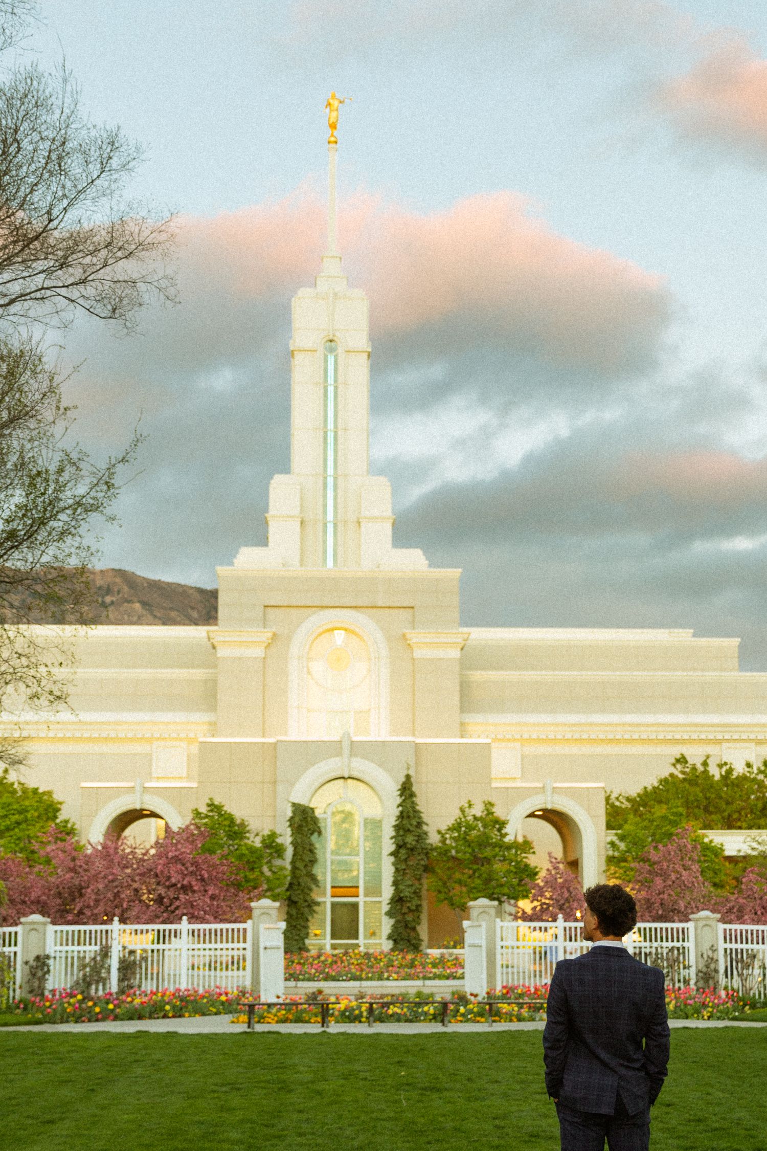 Person standing before an LDS Mormon Temple with golden spire, blooming trees, and dramatic sky at dusk.