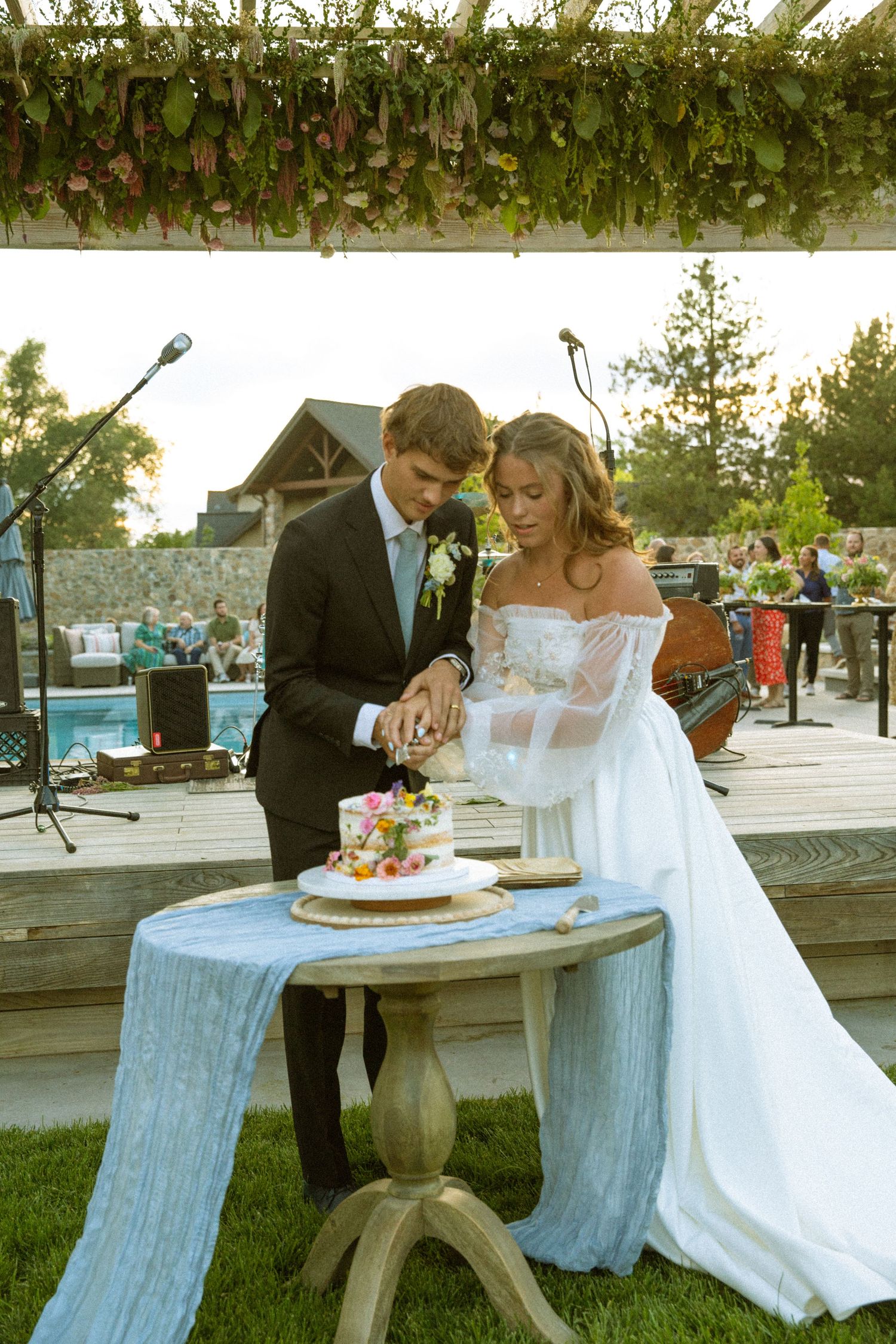 Newlyweds cut their wedding cake together at a rustic outdoor table.