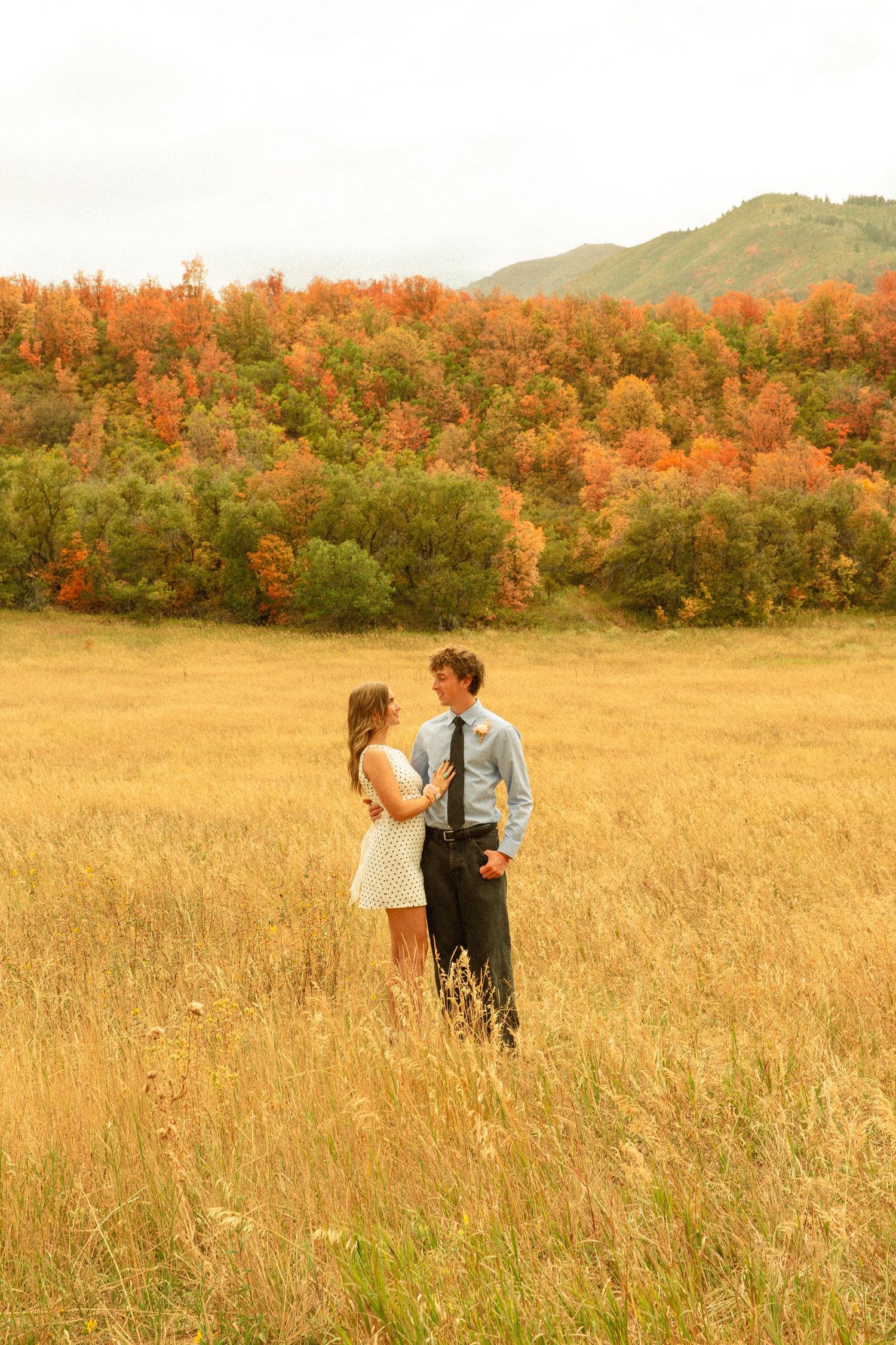 Two people stand together in a sunlit meadow with fall-colored trees lining the horizon.