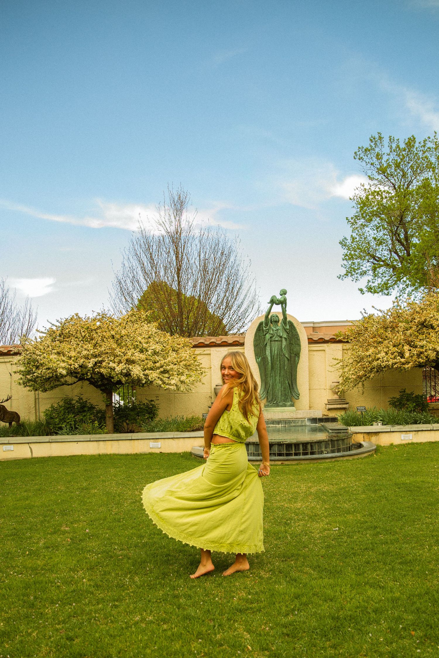 Woman in yellow dress twirling on green lawn near bronze statue fountain with blooming trees under blue sky.