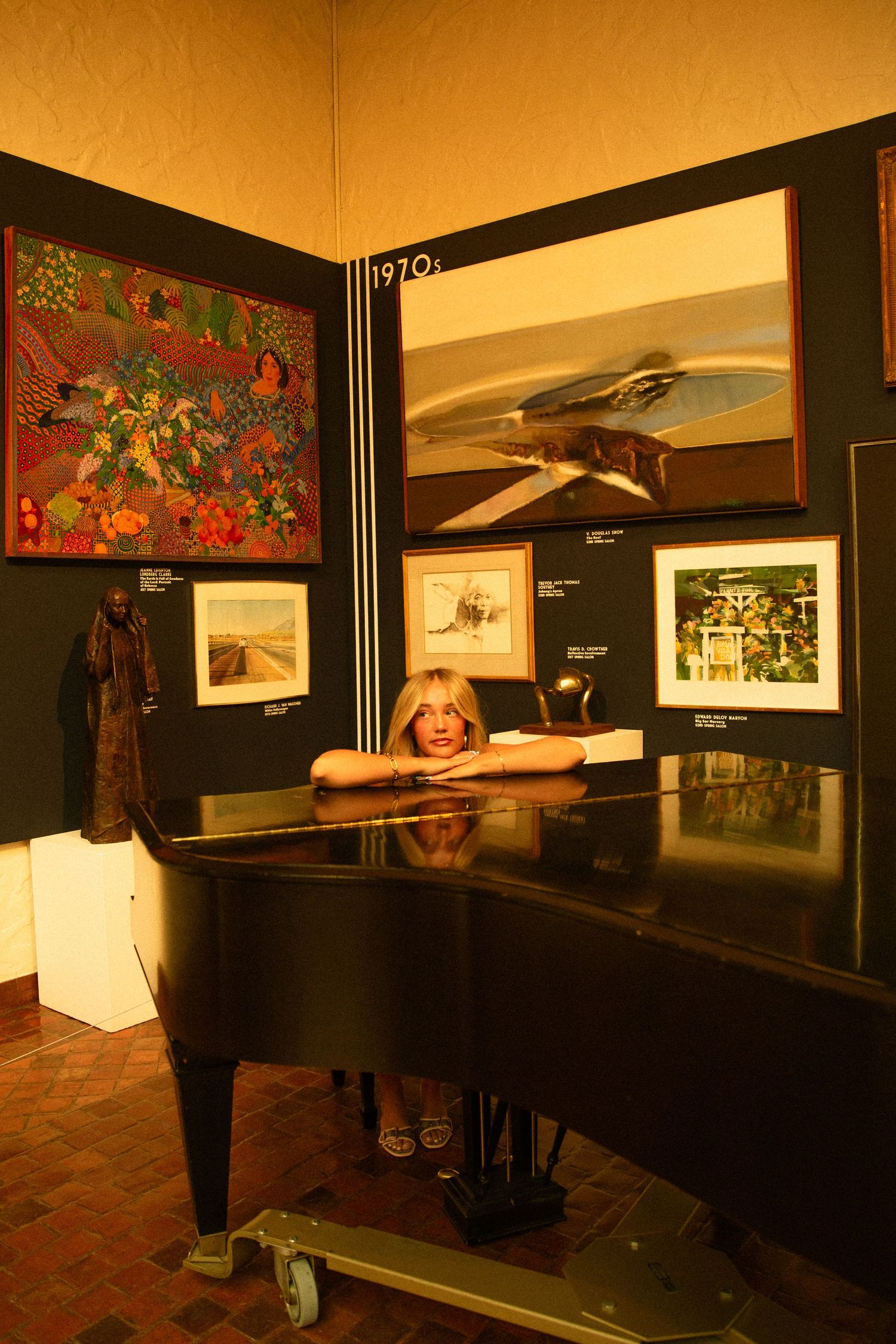 Woman posing behind a black grand piano in an art gallery with colorful paintings on dark walls.