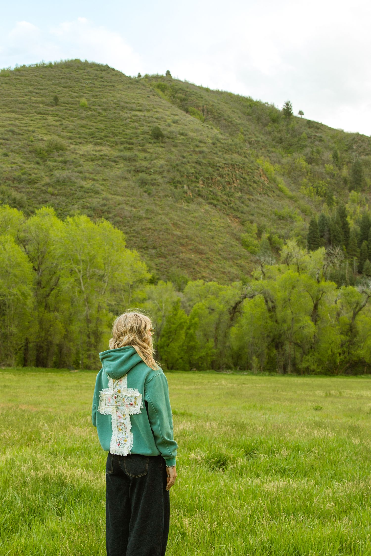 Blonde woman in green cross hoodie stands in lush meadow facing a tree-covered mountain landscape.