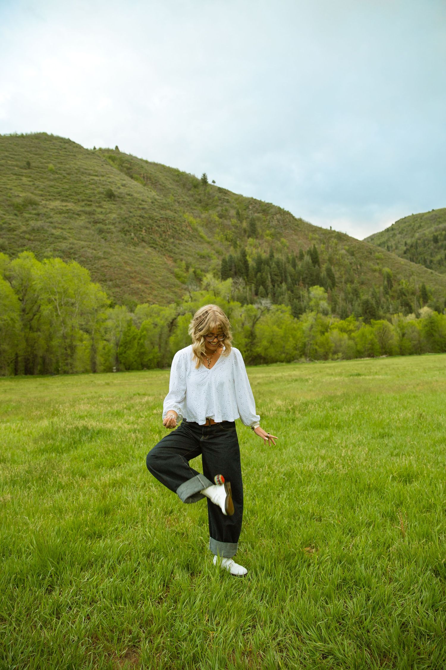 Woman in white blouse and black pants standing in green meadow with tree-covered hills in background.