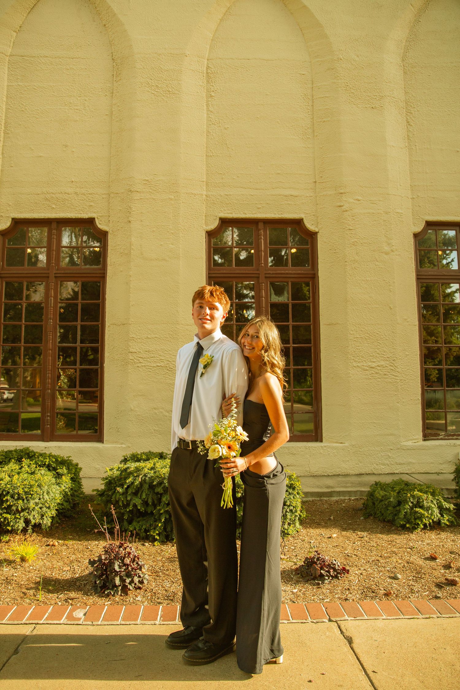 A young couple in formal attire poses outside a white building, the girl holding a yellow bouquet while leaning on her date.