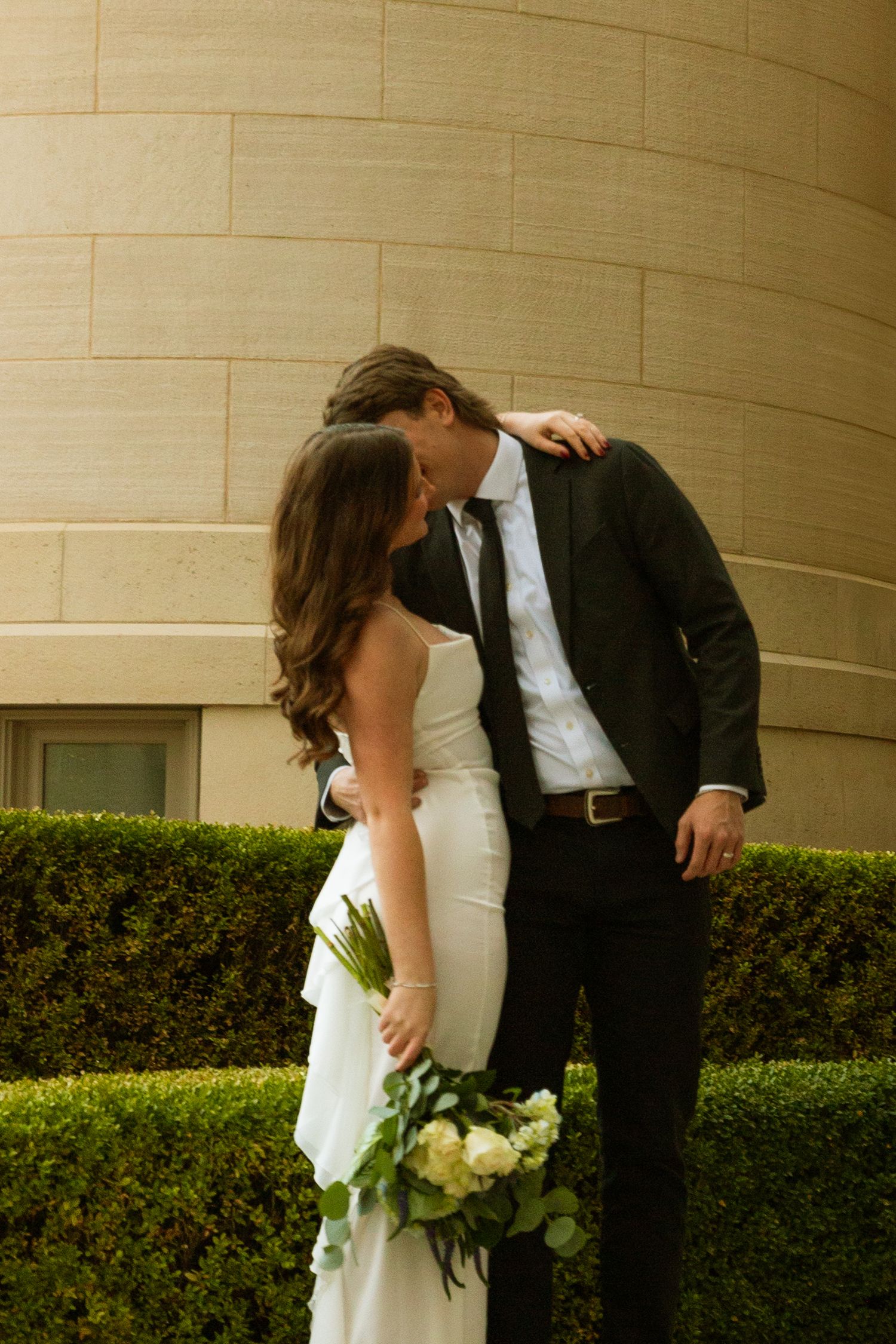 Romantic wedding couple shares intimate moment against beige wall with manicured hedges in outdoor garden setting.