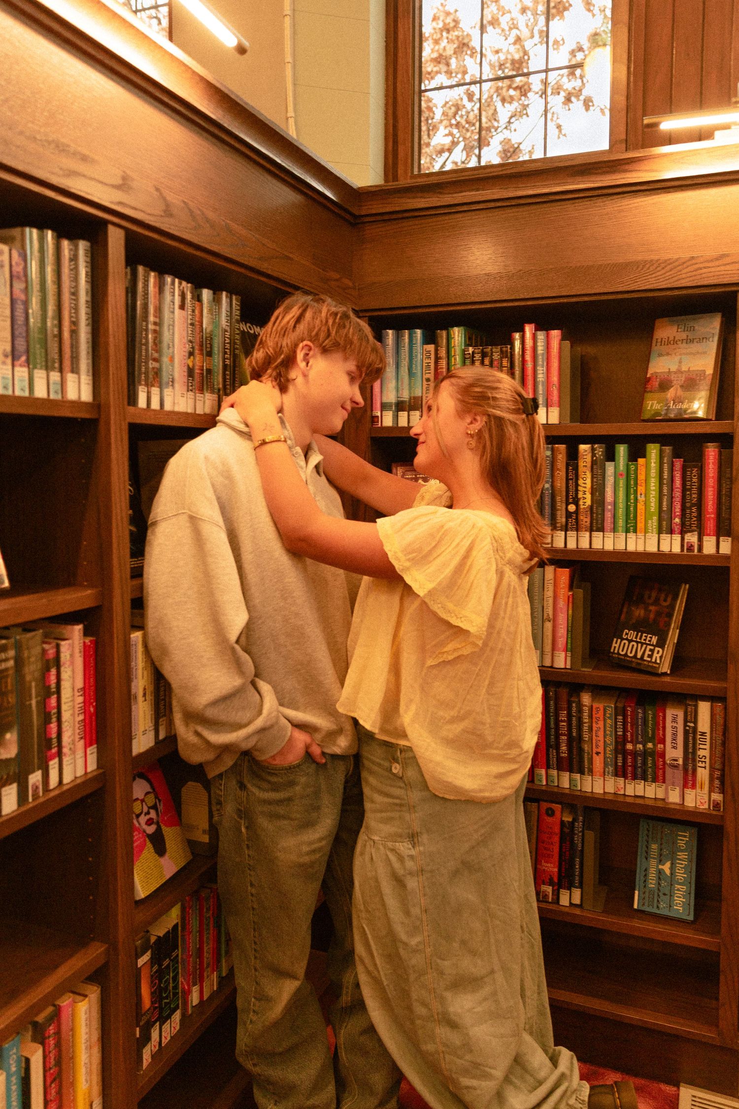 Two people share an intimate moment between library bookshelves with warm lighting coming through a window above.