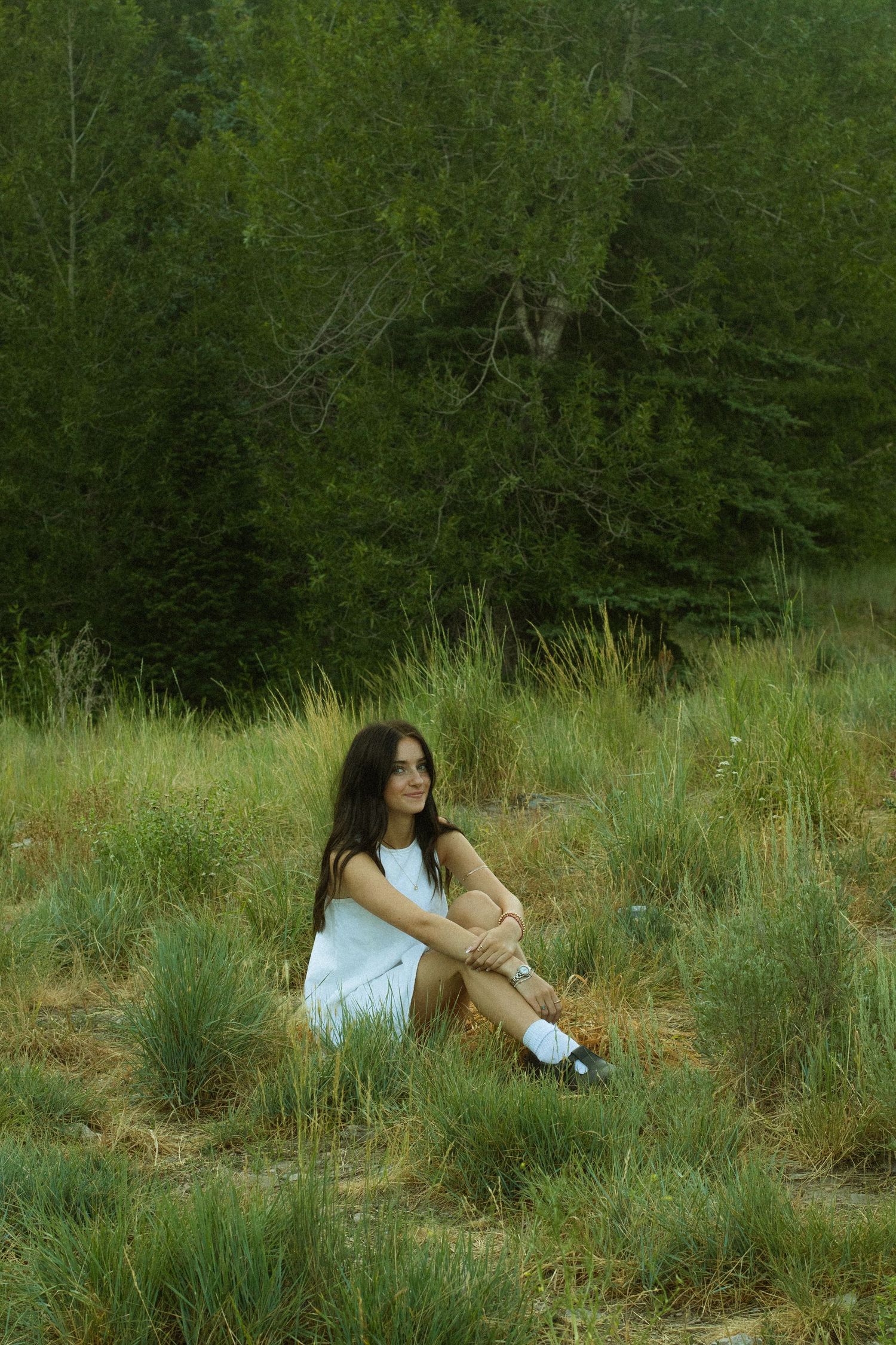 A person in a white top sits thoughtfully in tall grass surrounded by lush green trees and natural scenery.