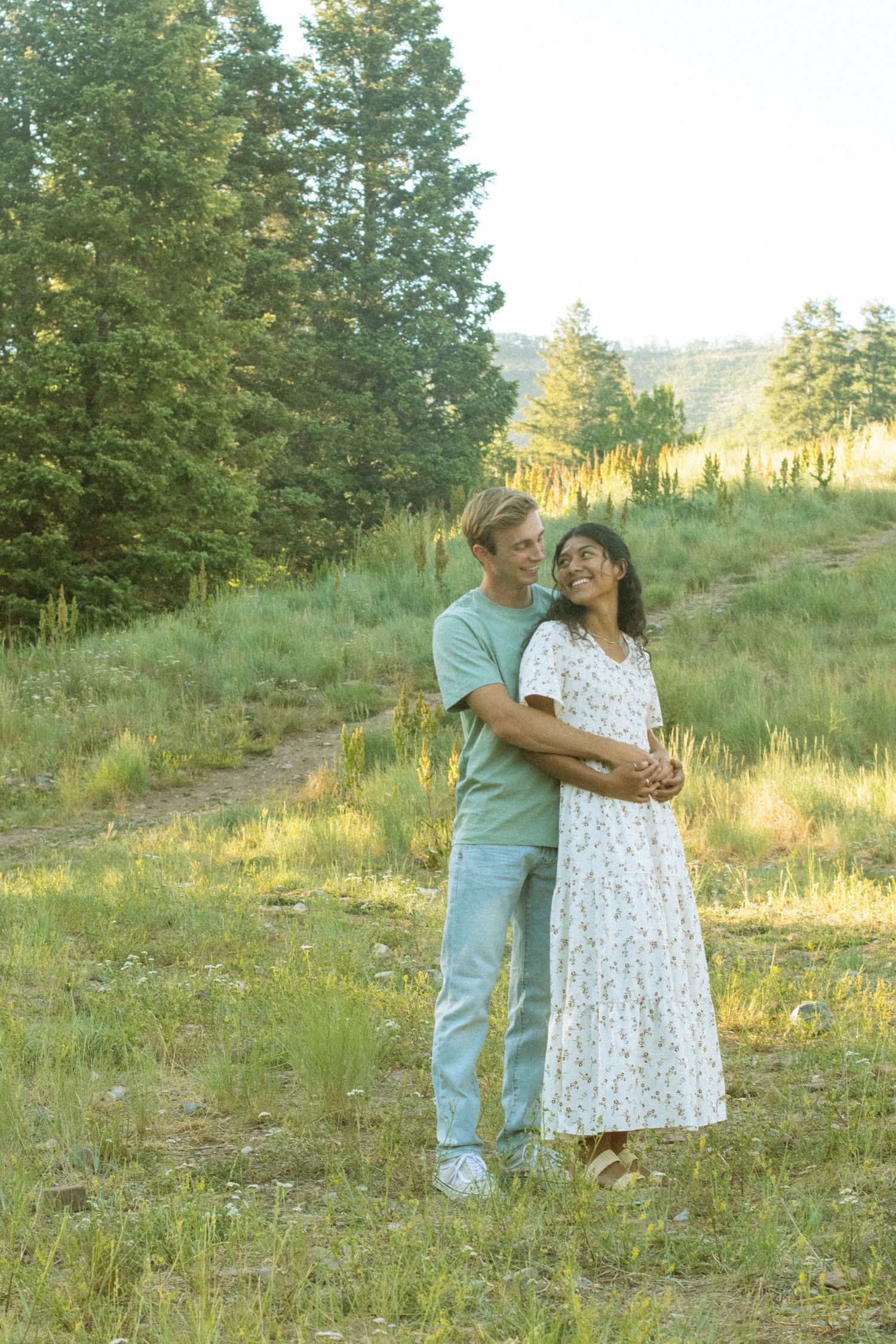 A couple shares a romantic moment together on a grassy hillside with evergreen trees in the background at sunset.