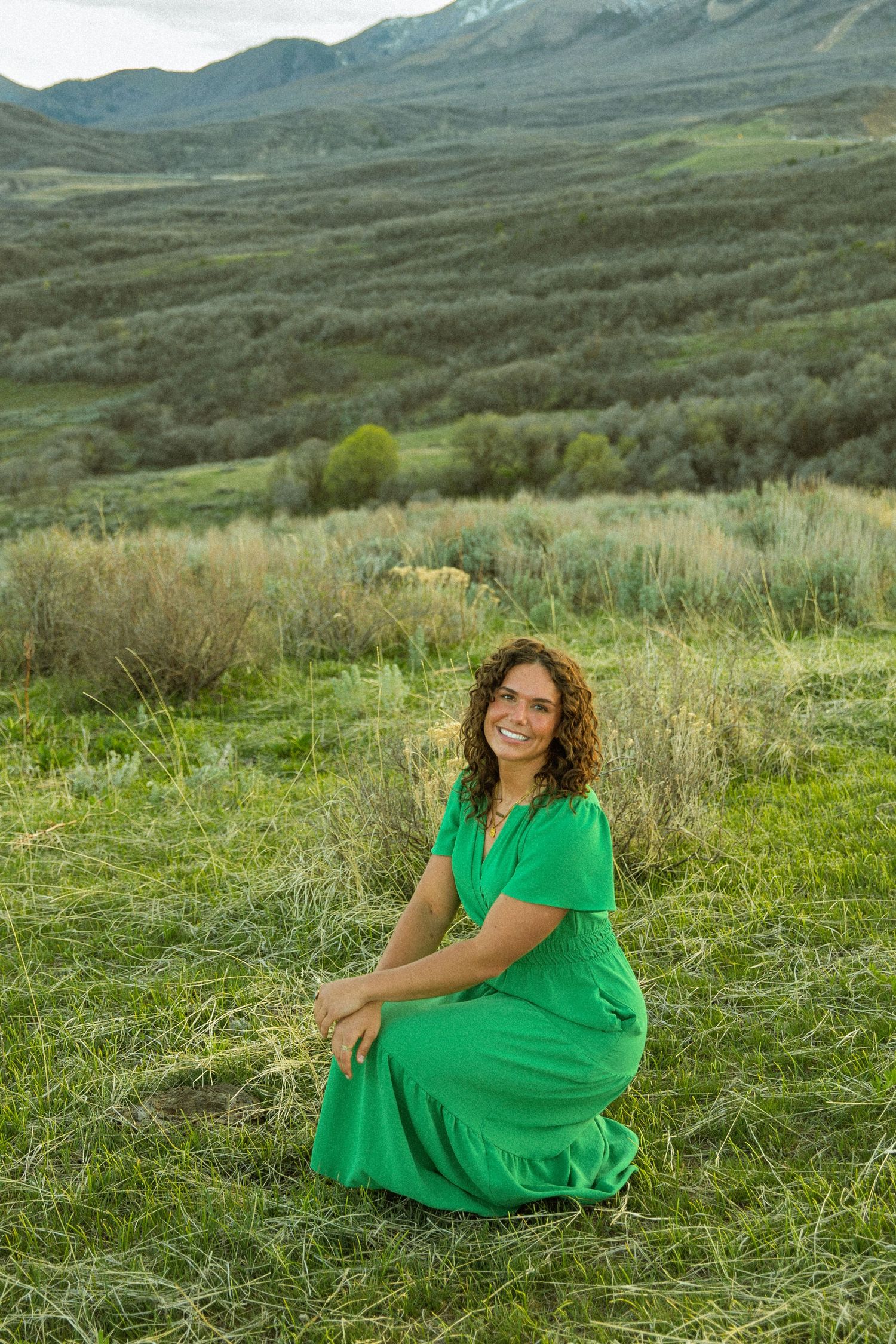 Woman in green dress sitting on grass with scenic mountain valley backdrop.