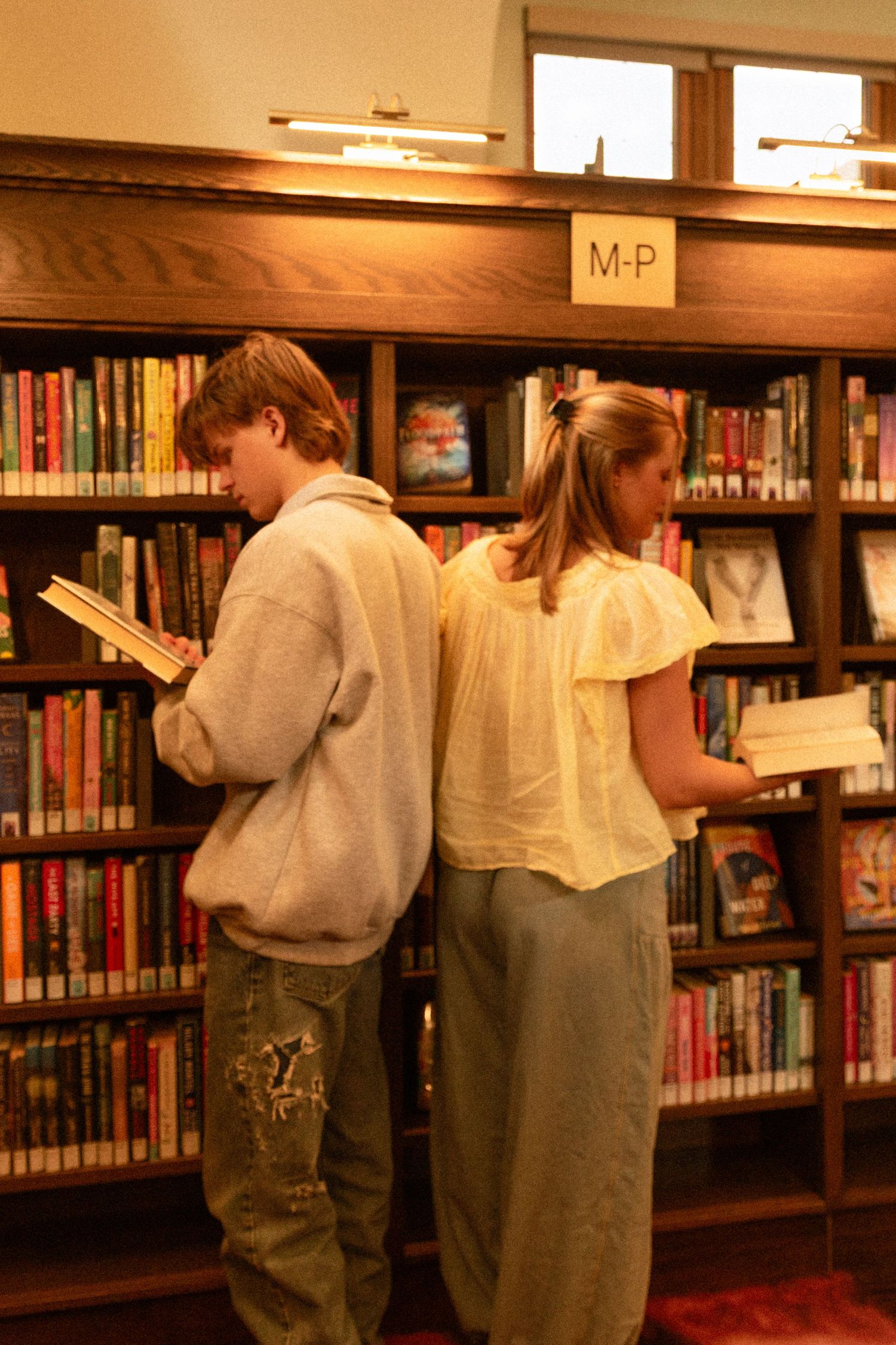 Two people stand back-to-back reading books in a library's M-P section with wooden shelves and warm lighting.