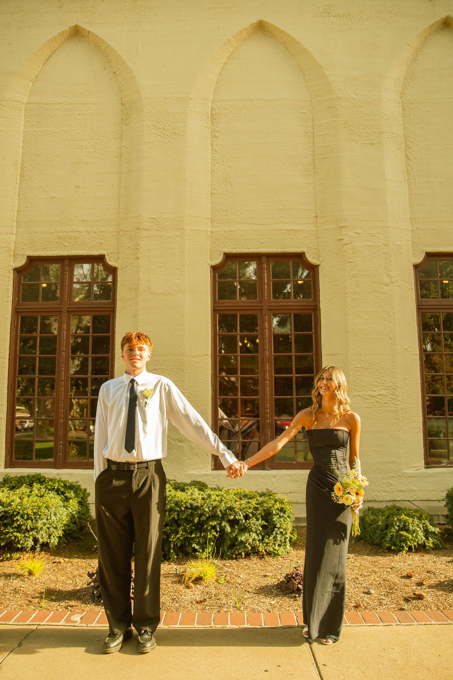 A young couple in formal attire holds hands outside a building, she in a black gown with flowers, he in a white shirt and tie.