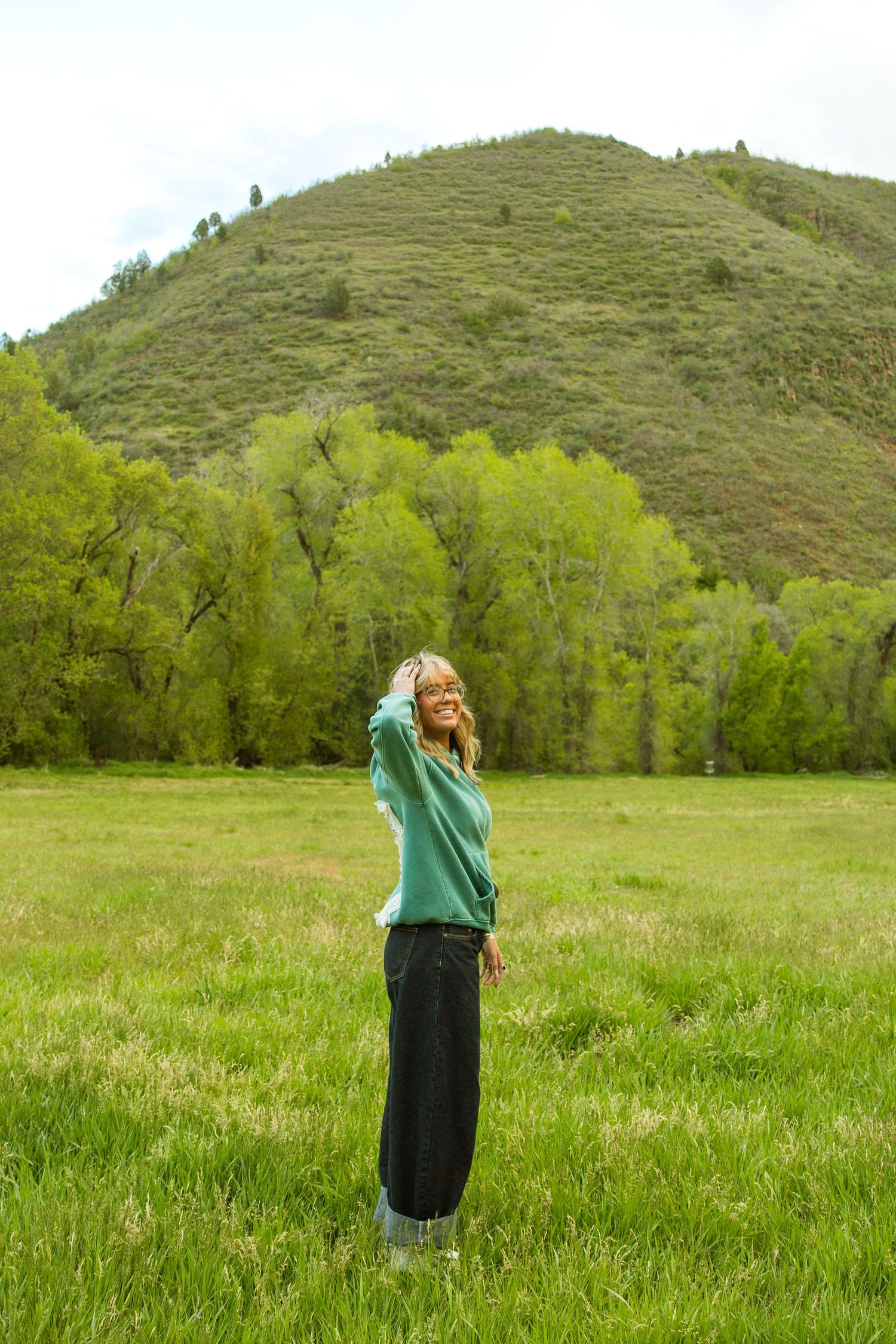 A smiling woman in a teal sweater and black pants stands in a lush green meadow with tree-covered hills behind her.