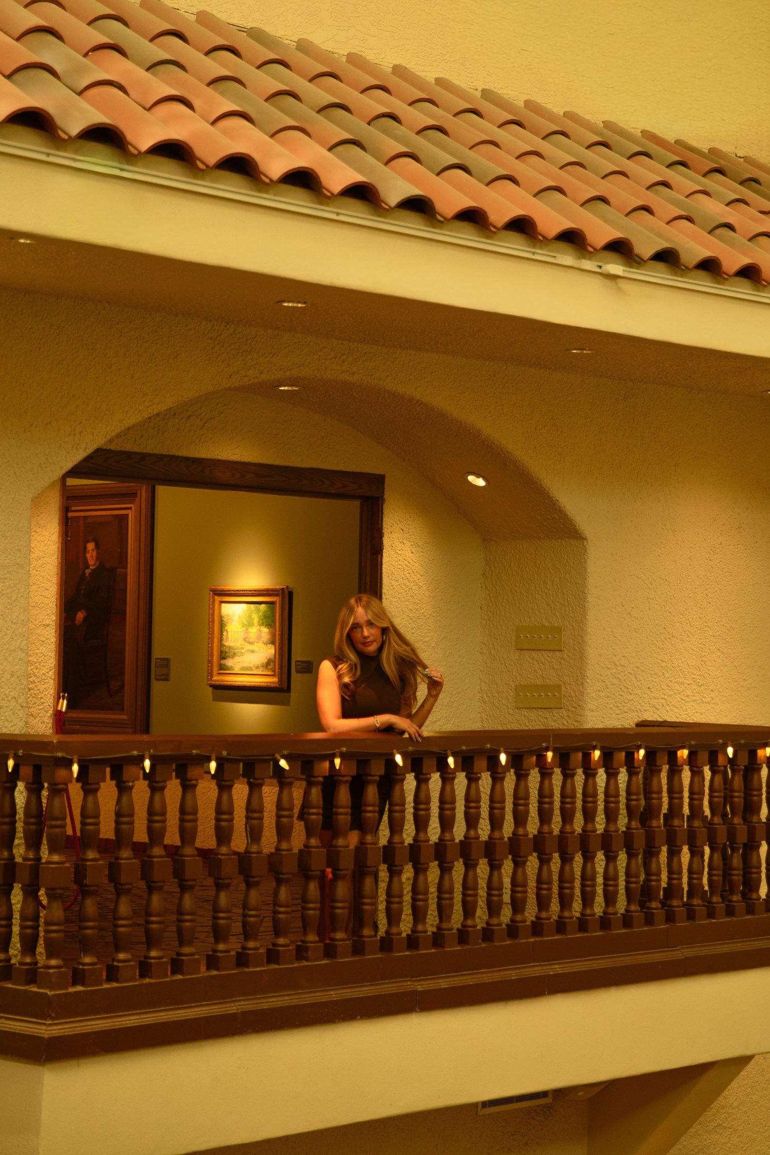 Woman leaning on ornate wooden balcony railing in elegant Spanish-style building with arched doorway and artwork.