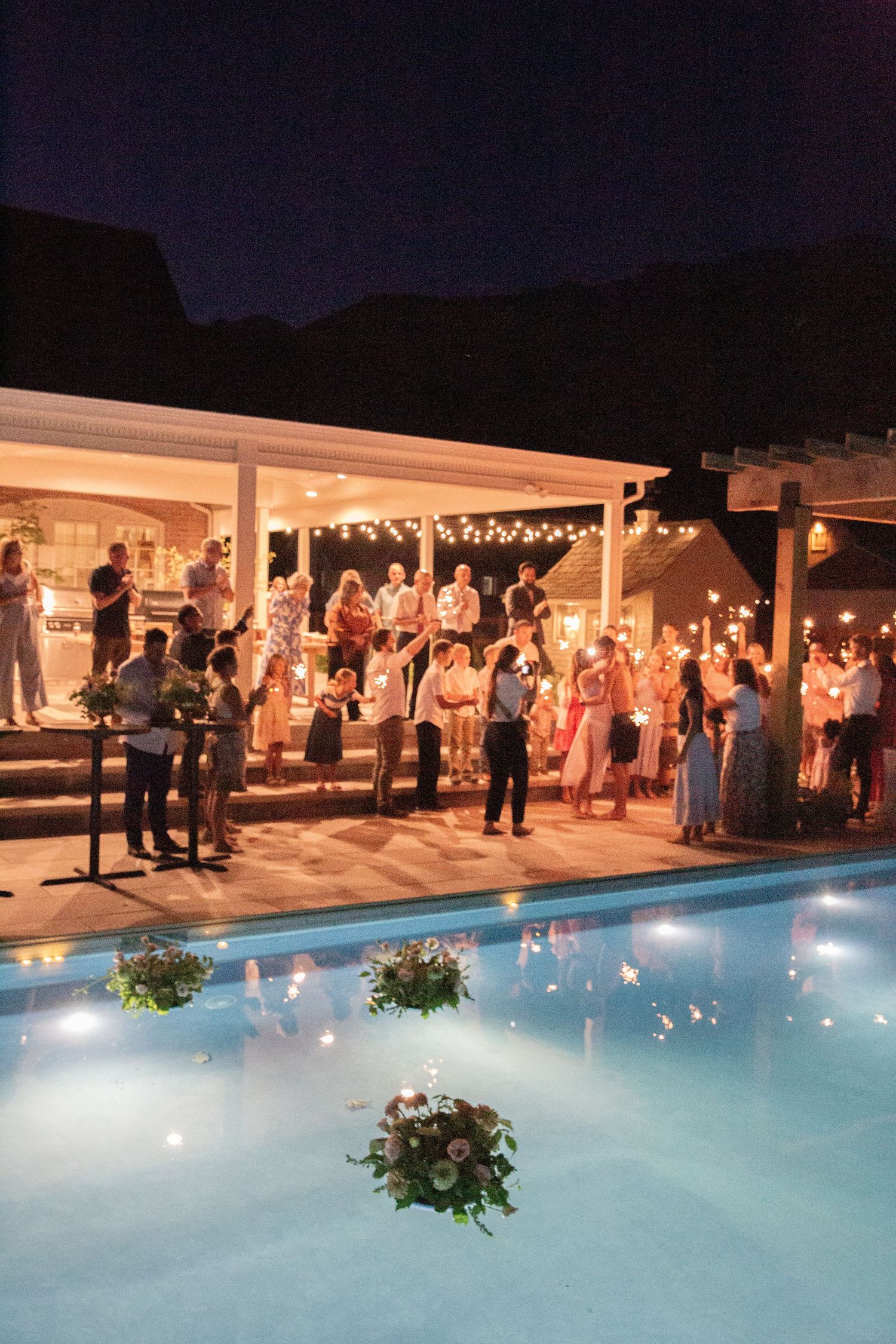 Nighttime poolside reception with string lights illuminating guests gathered along a white-columned venue.