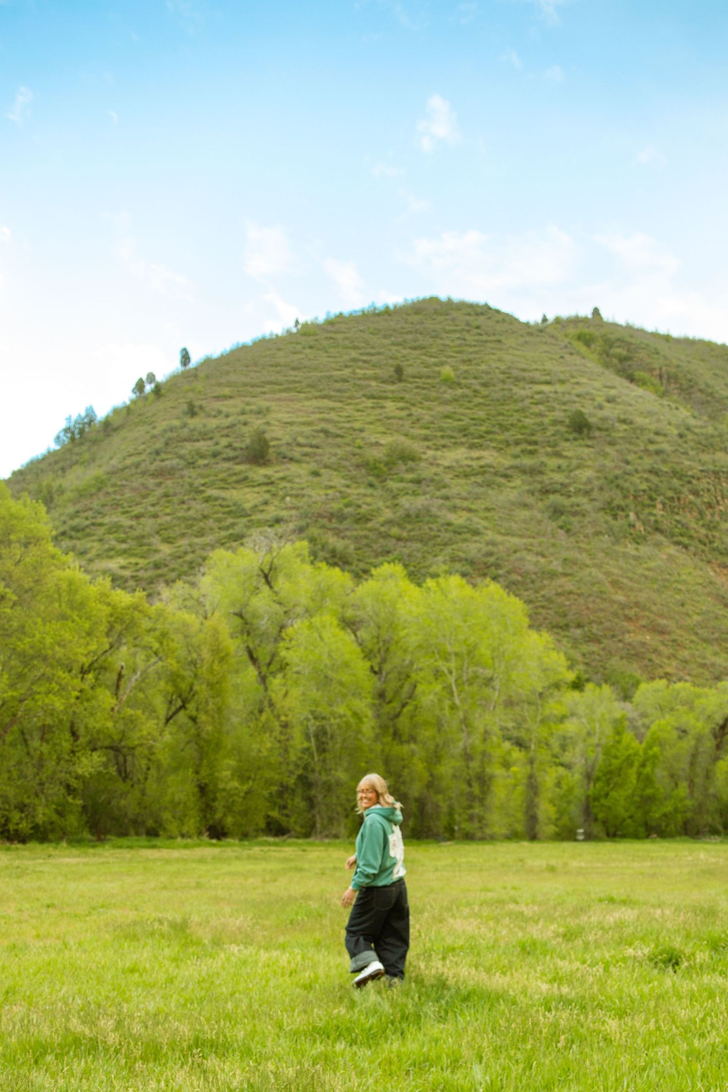 A person stands in a lush green meadow with a tree-covered hill and blue sky in the background.