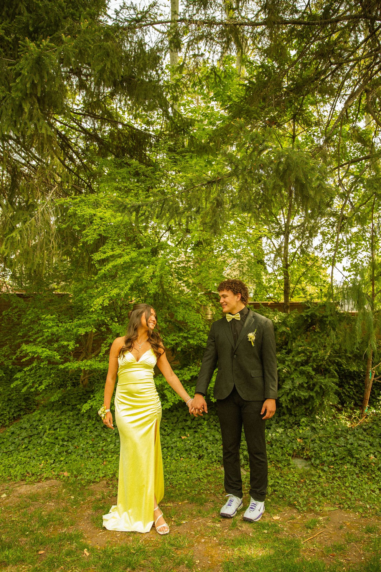 A couple in formal attire holds hands outdoors, she in a yellow gown, he in a dark suit with boutonniere.