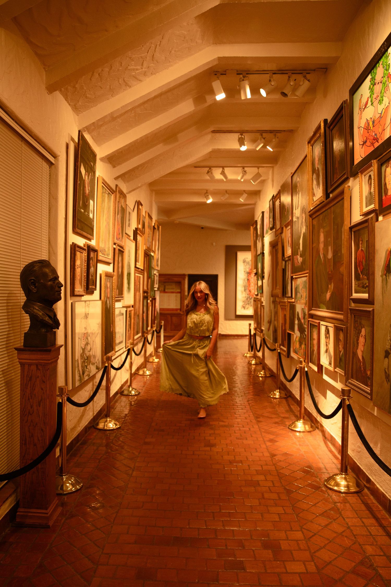 A woman in a green dress walks through a warmly lit art gallery corridor lined with paintings and a bronze bust sculpture.