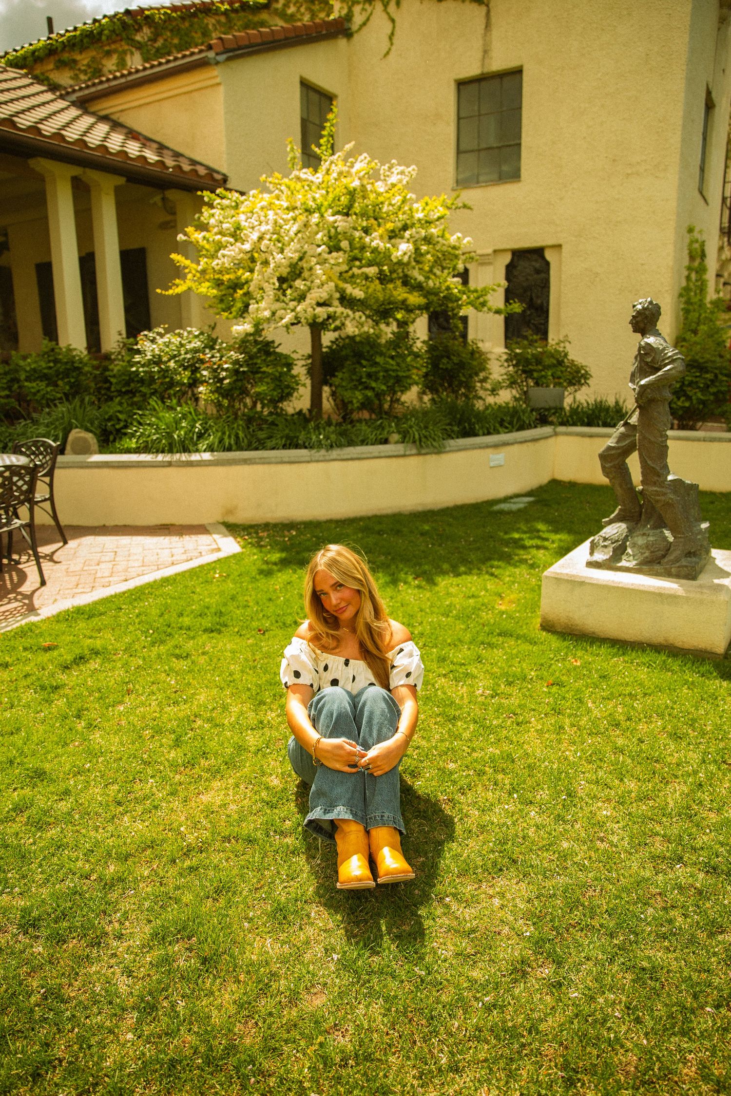 Young woman in white top and jeans sits cross-legged on lush green lawn beside elegant building with statue and flowering tree.