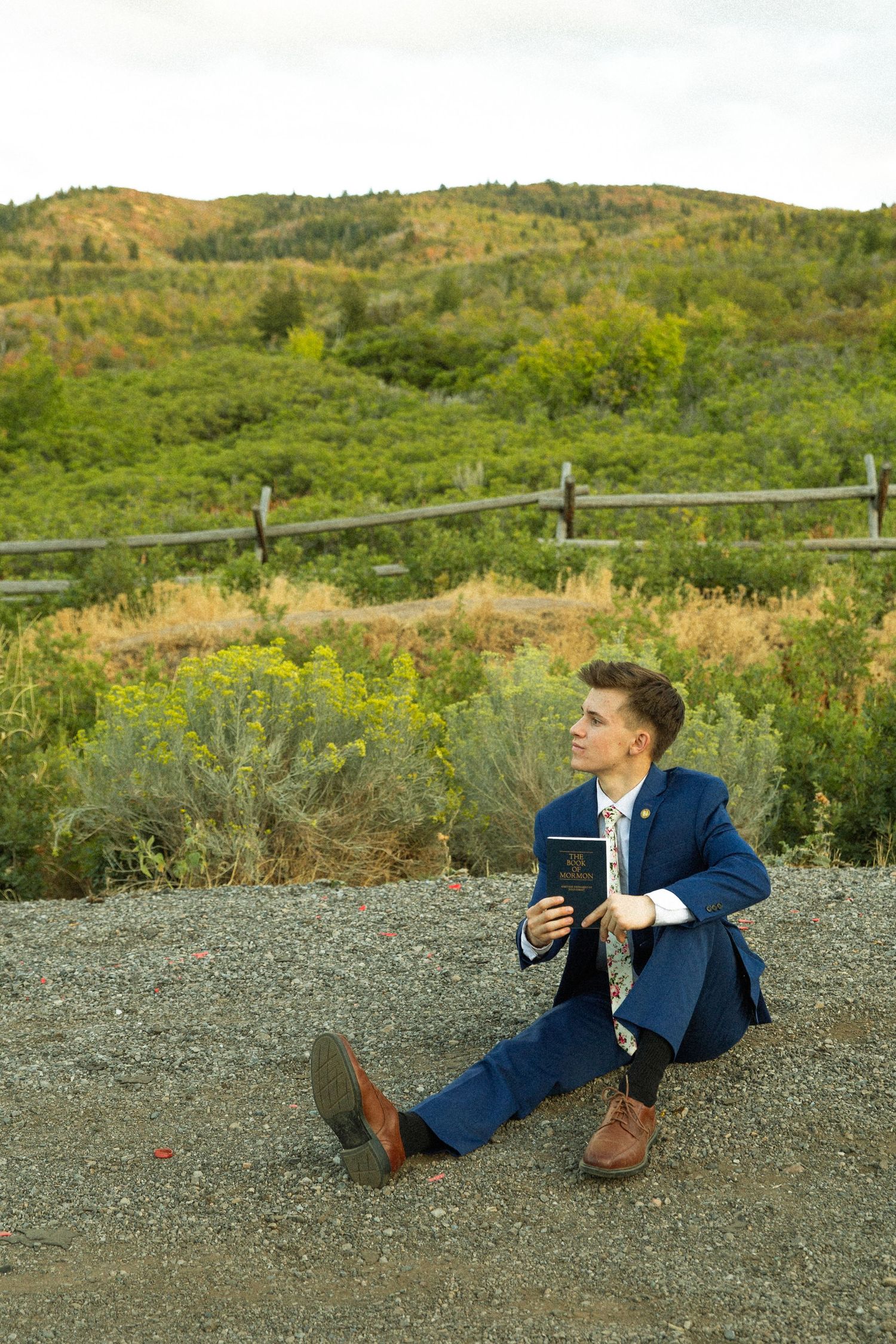A person in a navy blue suit lies on gravel ground while taking photos in a scenic mountain landscape.