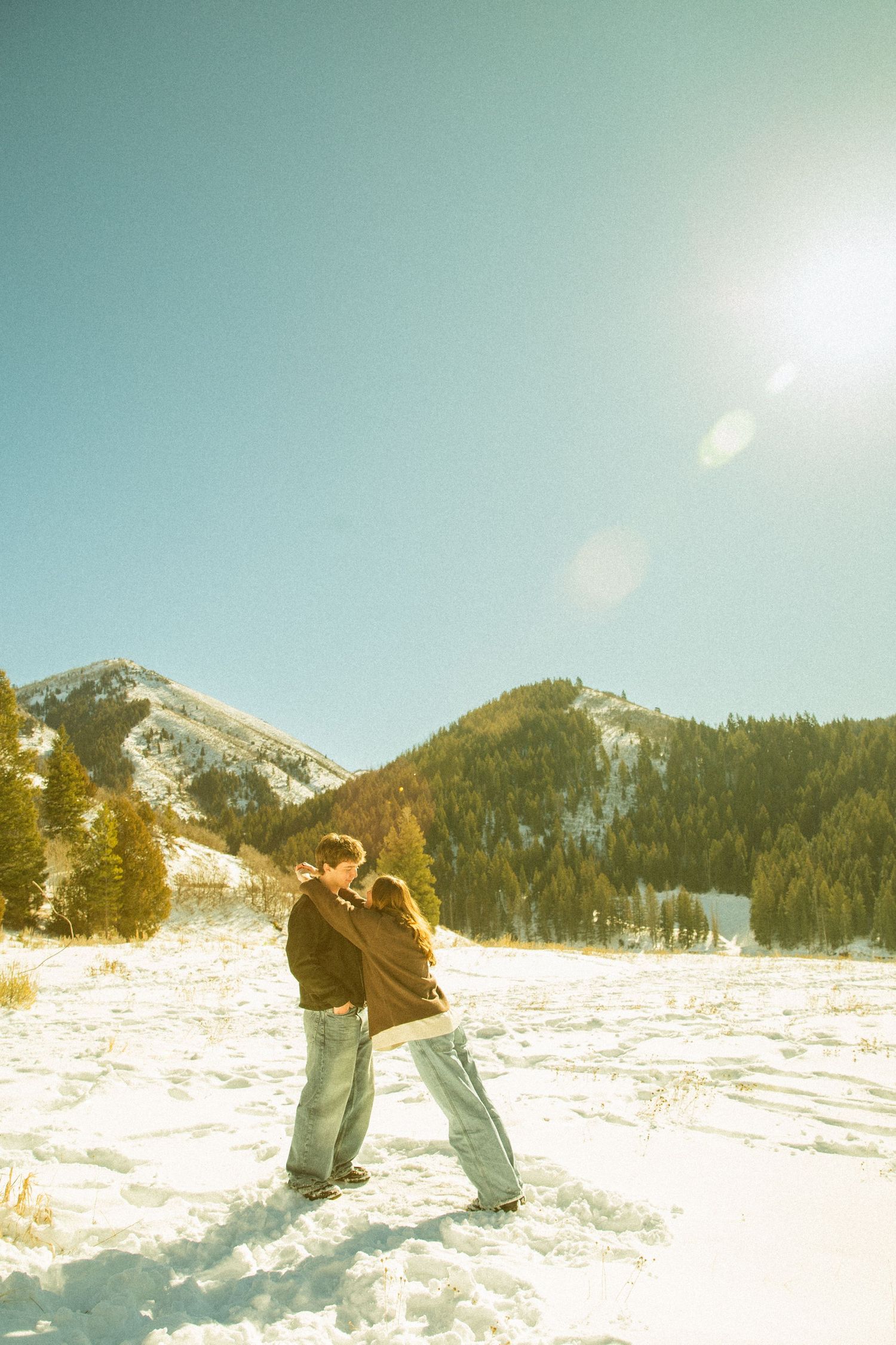 A couple embraces while standing in deep snow with mountain peaks and evergreen forest in the background.