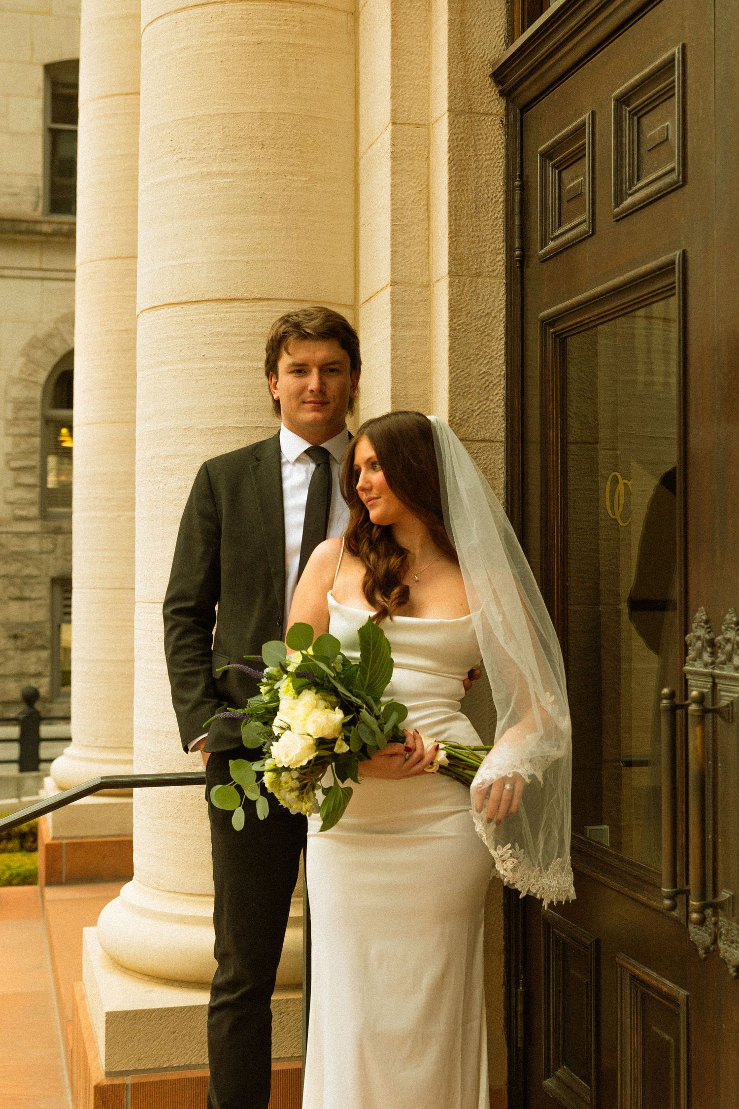 A wedding couple poses together against a grand column outside a historic building at sunset.