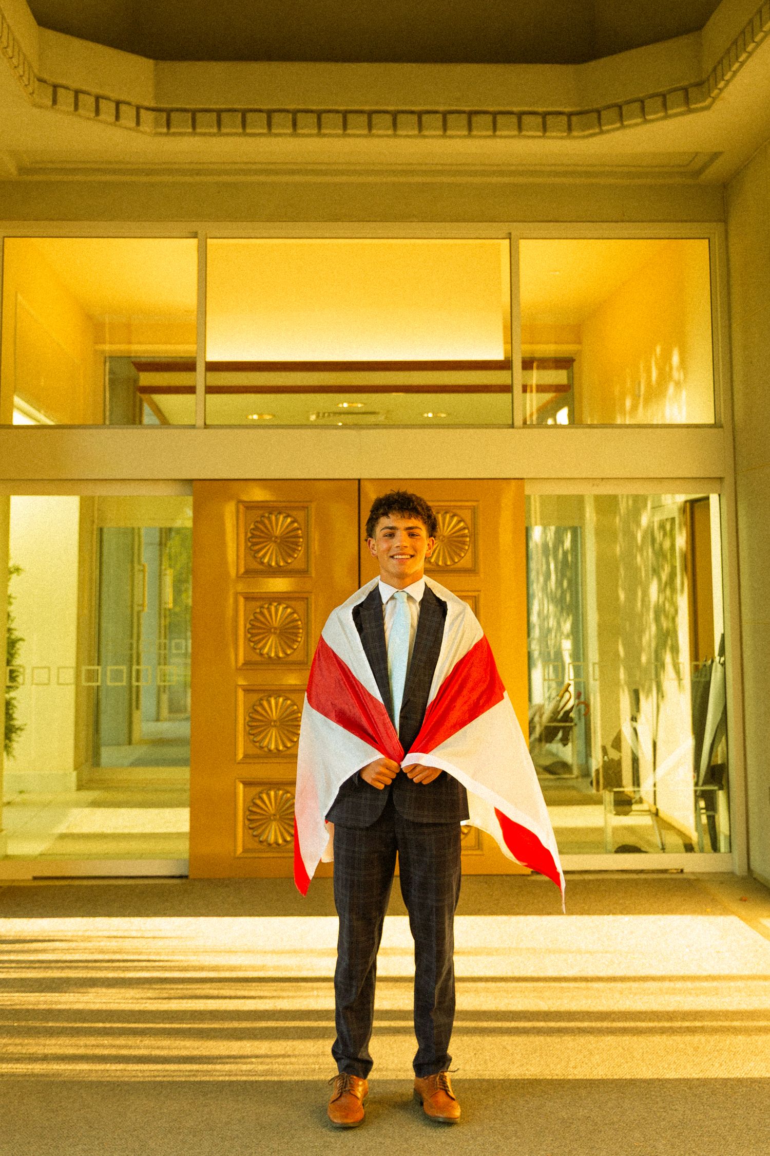 Young man in suit holding a red and white flag, standing before an illuminated building entrance at night.