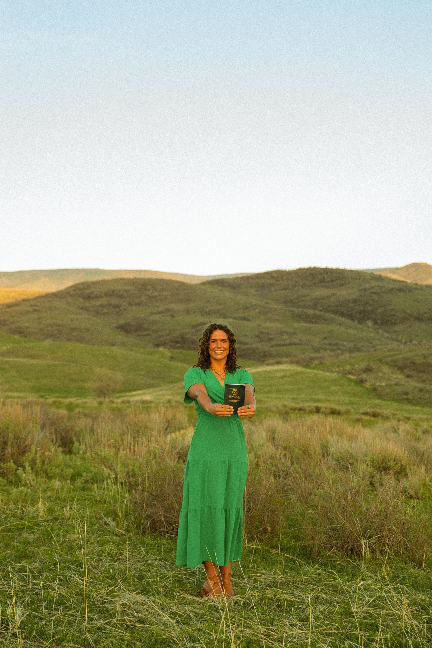 Woman in green dress holding a book stands in lush green rolling hills landscape at golden hour.